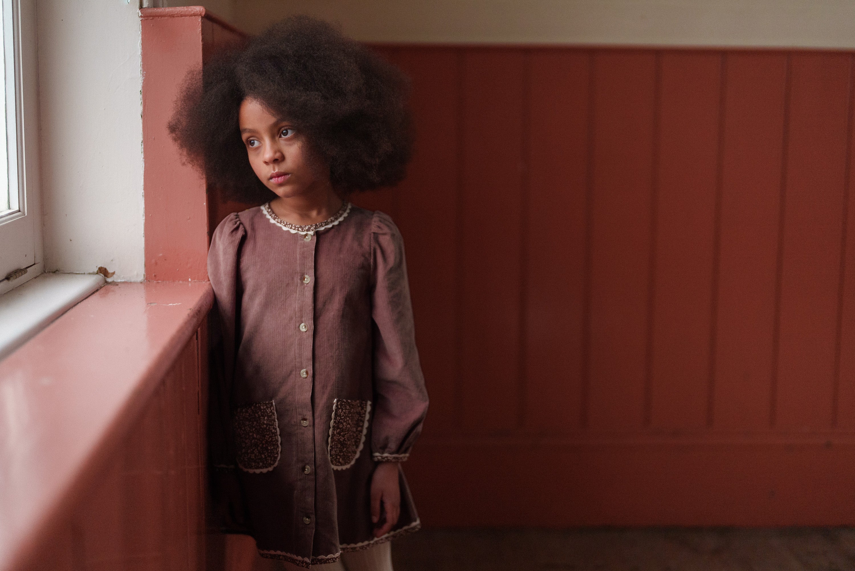 Young girl wearing a nutmeg-colored velvet dress with floral pocket details, made from sustainable organic cotton, embodying vintage-inspired children's clothing.