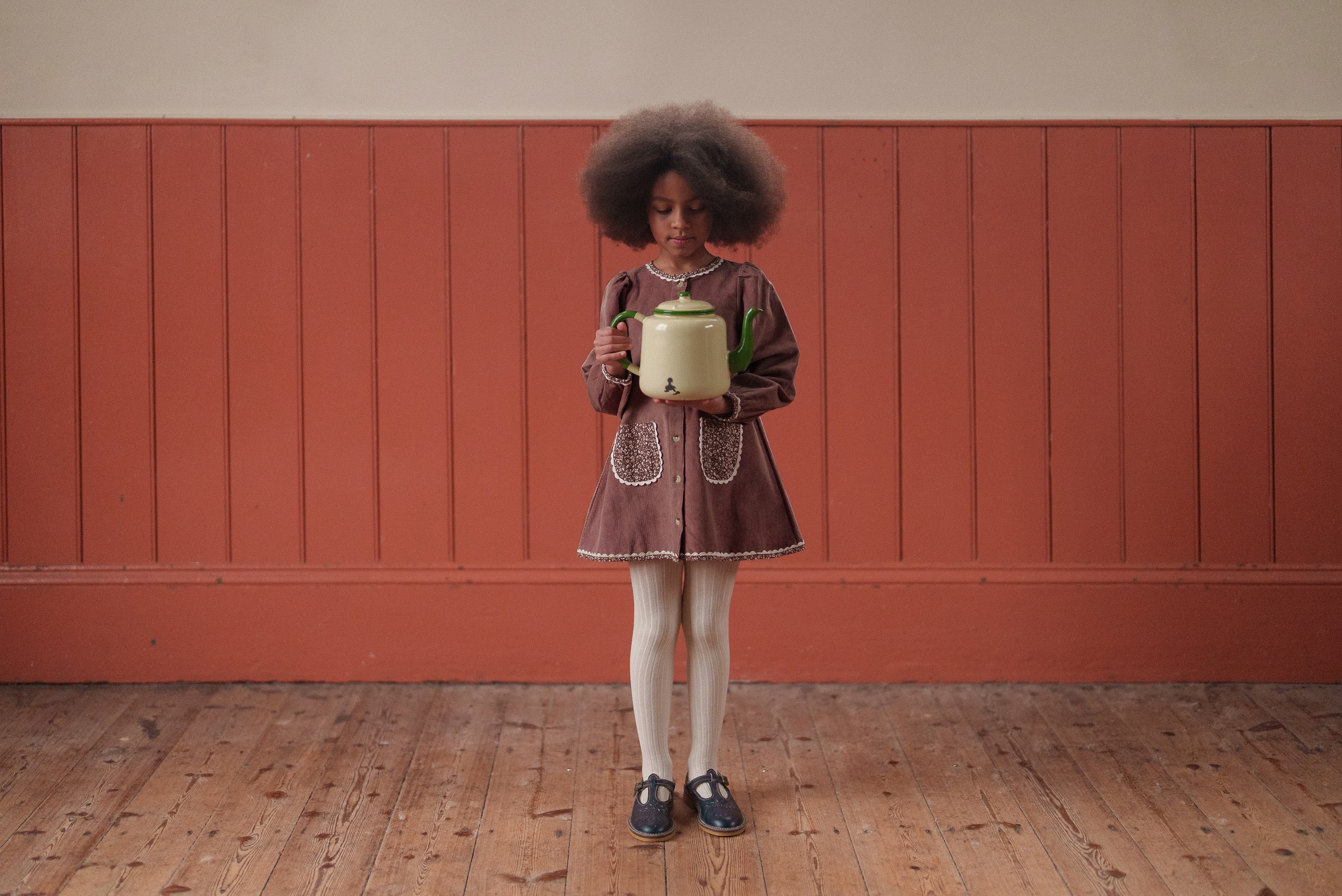 Young girl wearing a vintage style dress in organic cotton and velvet in a warm nutmeg color, standing on wooden floor against a muted red wooden panel wall, holding a green pot.