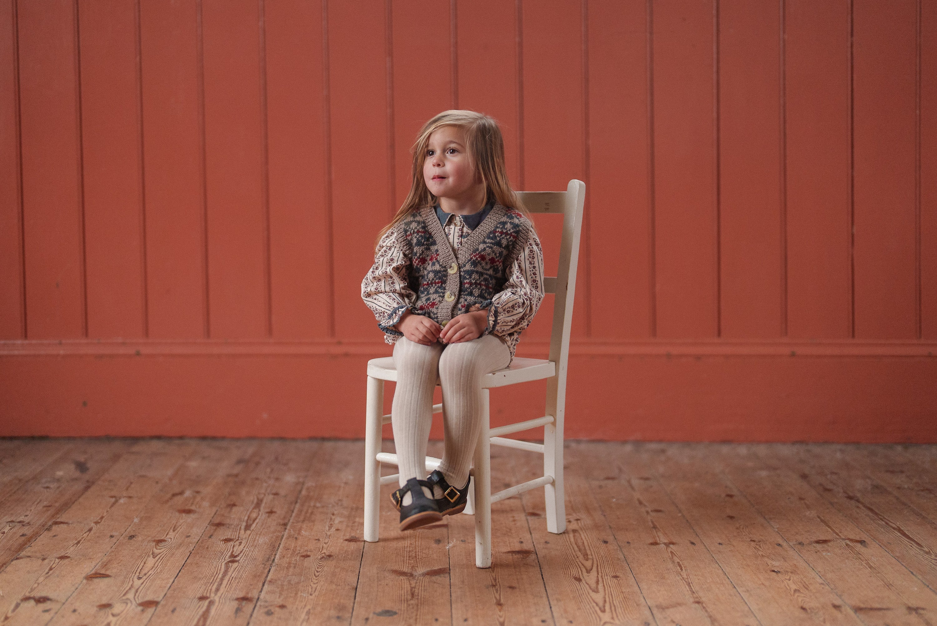 Young child wearing a hand-knitted multicolor merino wool tank top vest with beige tights and navy shoes, sitting on a white chair against a warm orange wooden wall in a vintage style setting.