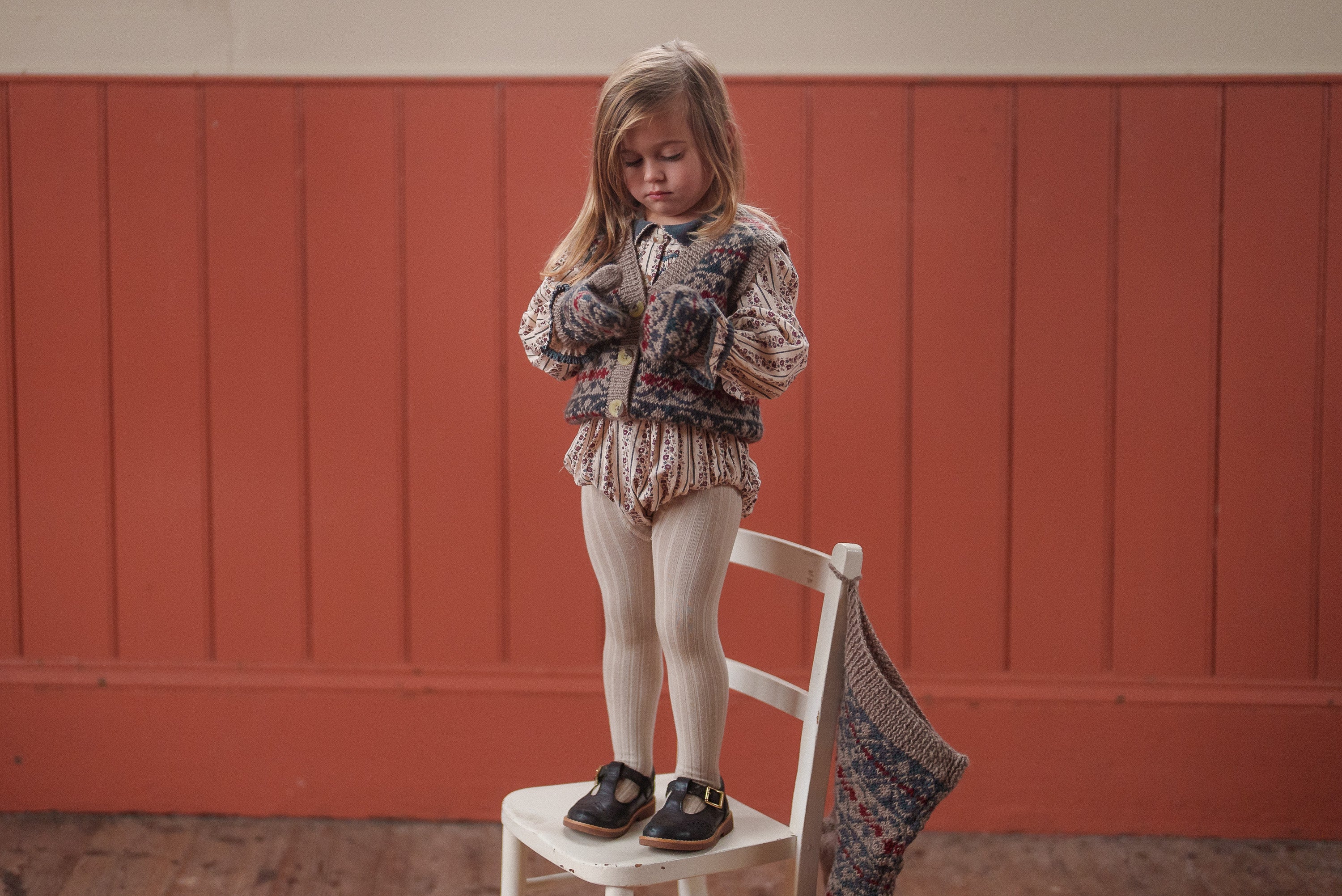 Young child wearing vintage style children's clothing in natural materials, including a hand-knitted fairisle merino vest, standing on a white chair in a warm, cozy setting.