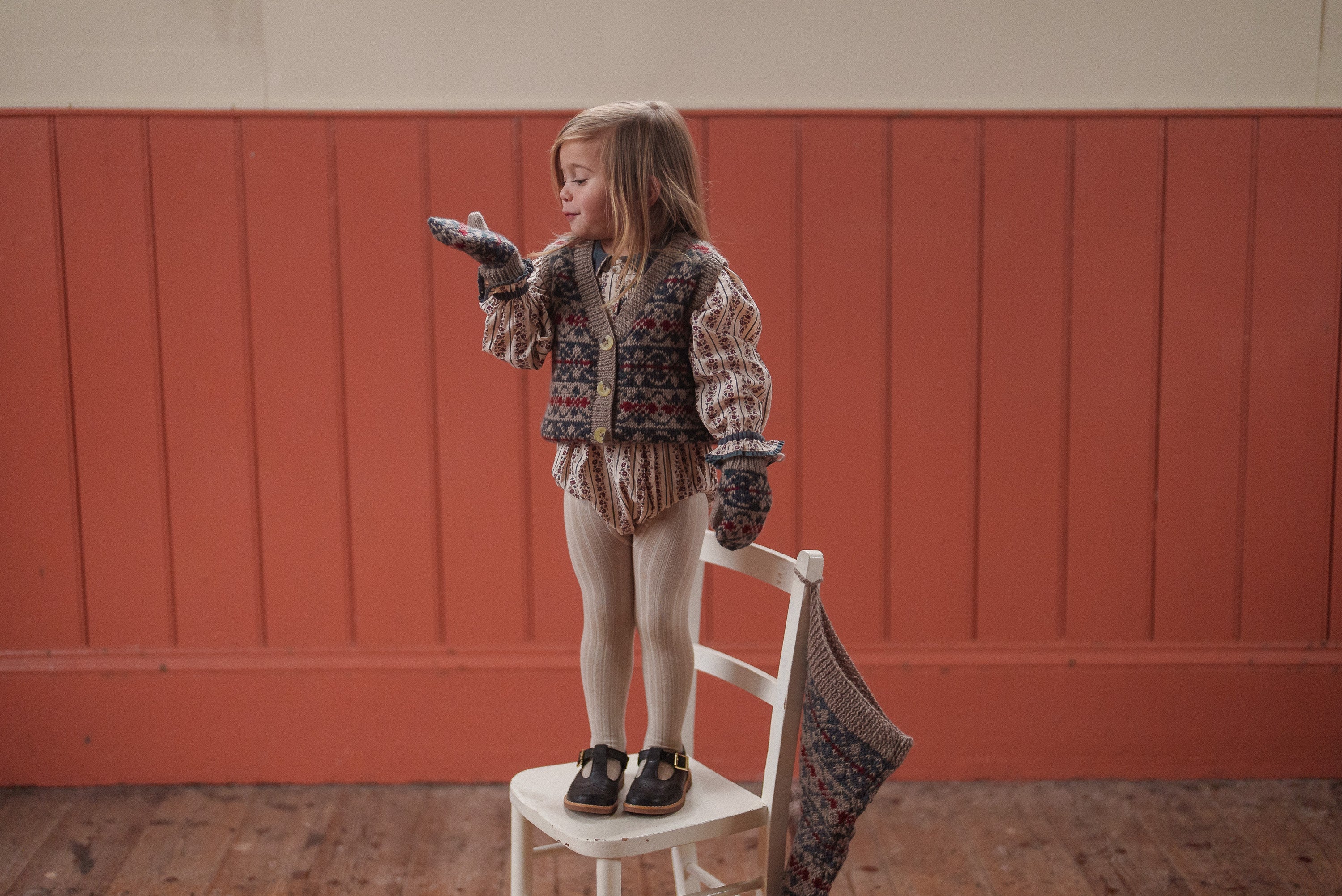 Young child wearing vintage style organic cotton clothing with knitted vest and mittens, standing on a white chair in a warm, rustic room.