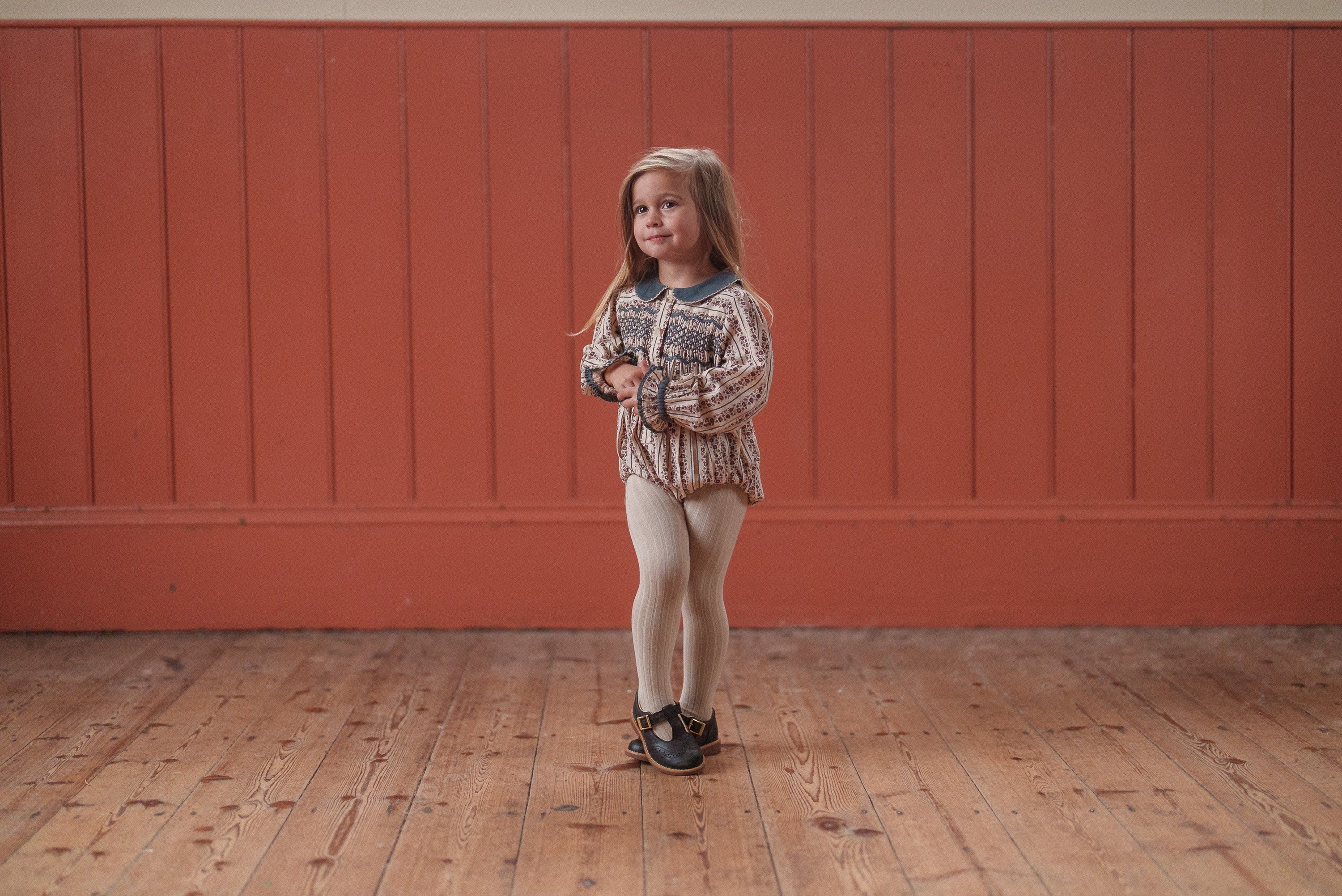 Young girl wearing a vintage-inspired organic cotton romper with a floral pattern, standing on a wooden floor against a warm-toned background, showcasing sustainable children's clothing.