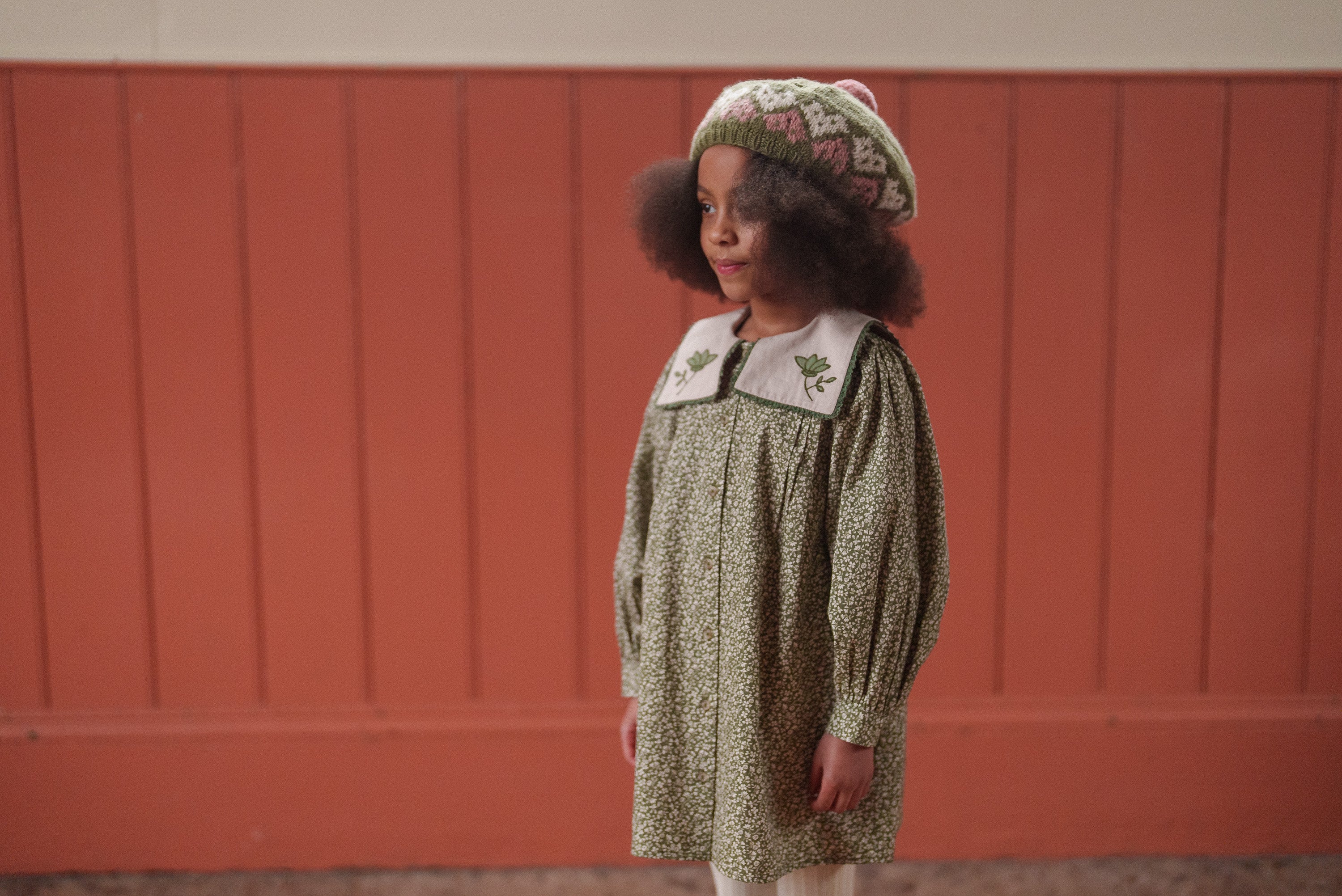 Young girl wearing a vintage style green floral organic cotton dress with embroidered collar and a knitted hat, showcasing sustainable and ethically made children's clothing.