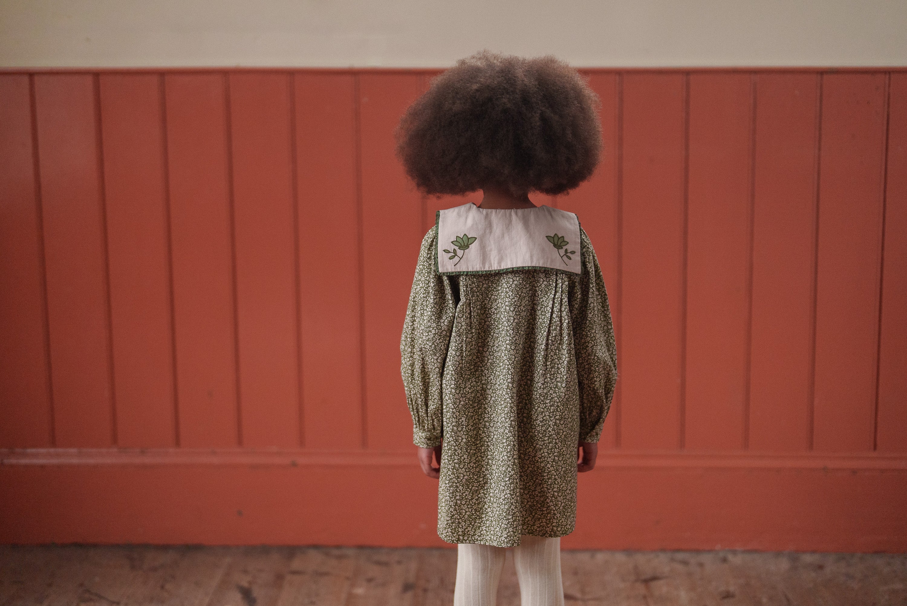 Child wearing a green organic cotton dress with a floral pattern and vintage-inspired collar, standing indoors against a warm wooden wall, showcasing sustainable children's clothing.