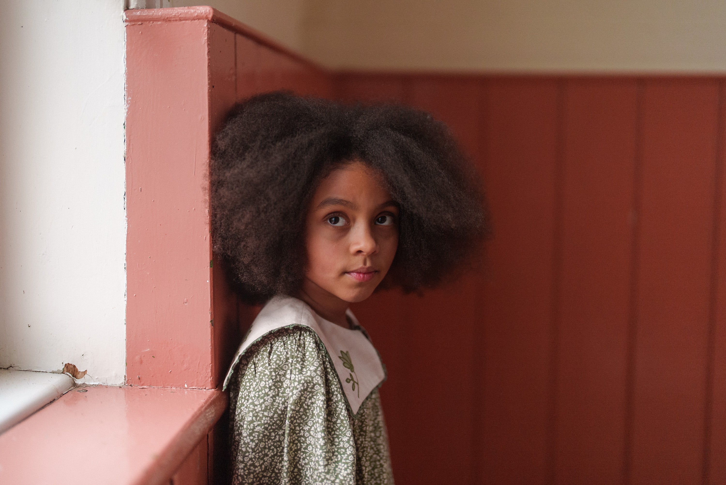 Young girl wearing a vintage-style green floral dress made from organic cotton, standing indoors with natural light highlighting her thoughtful expression.