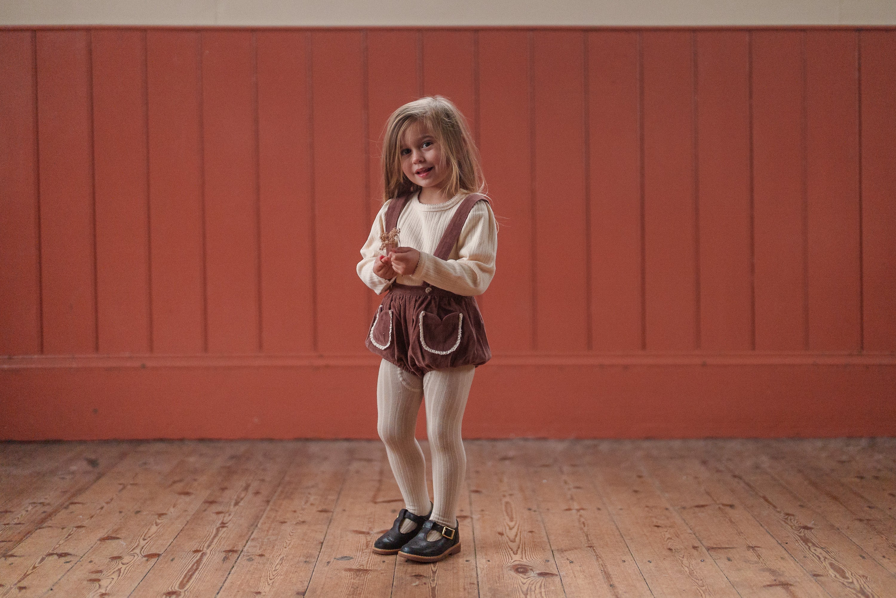 Toddler girl wearing a vintage-style baby romper in nutmeg velvet with organic cotton fabric, styled with white tights and black shoes against a warm wooden backdrop.