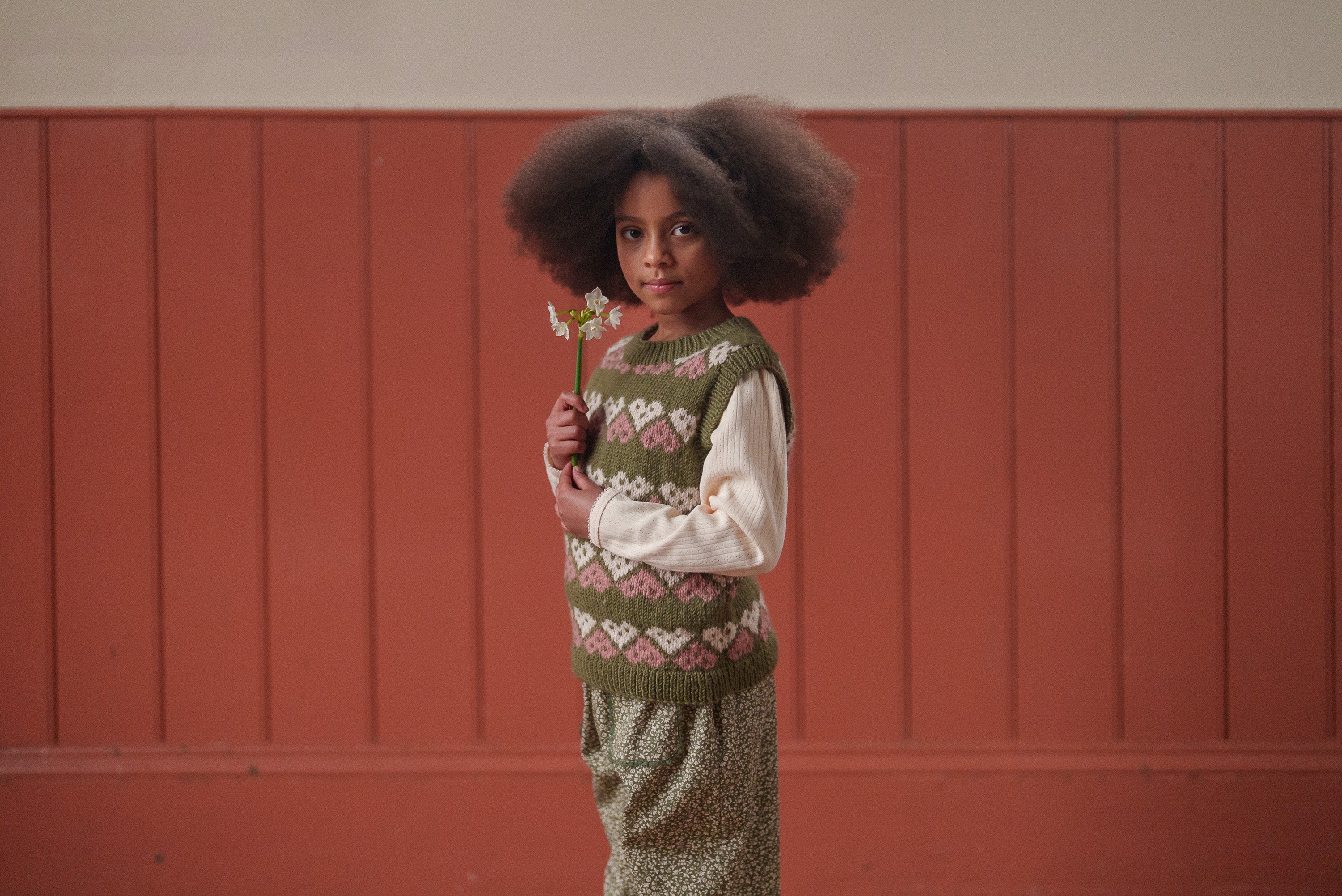 Child wearing a hand-knitted multicolor green heart tank top vest made from merino wool, paired with vintage-style sustainable kids clothing in an indoor setting.