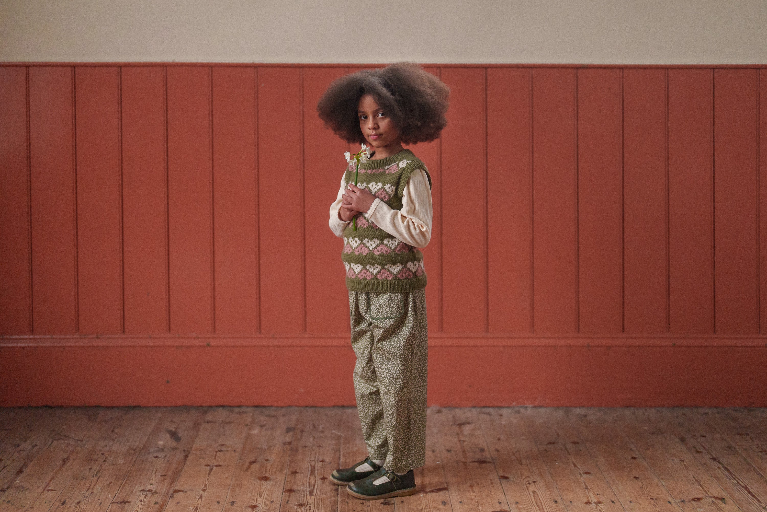 Child wearing vintage style multicolor hand-knitted heart tank top vest made from soft merino wool paired with patterned pants, showcasing sustainable children's clothing.