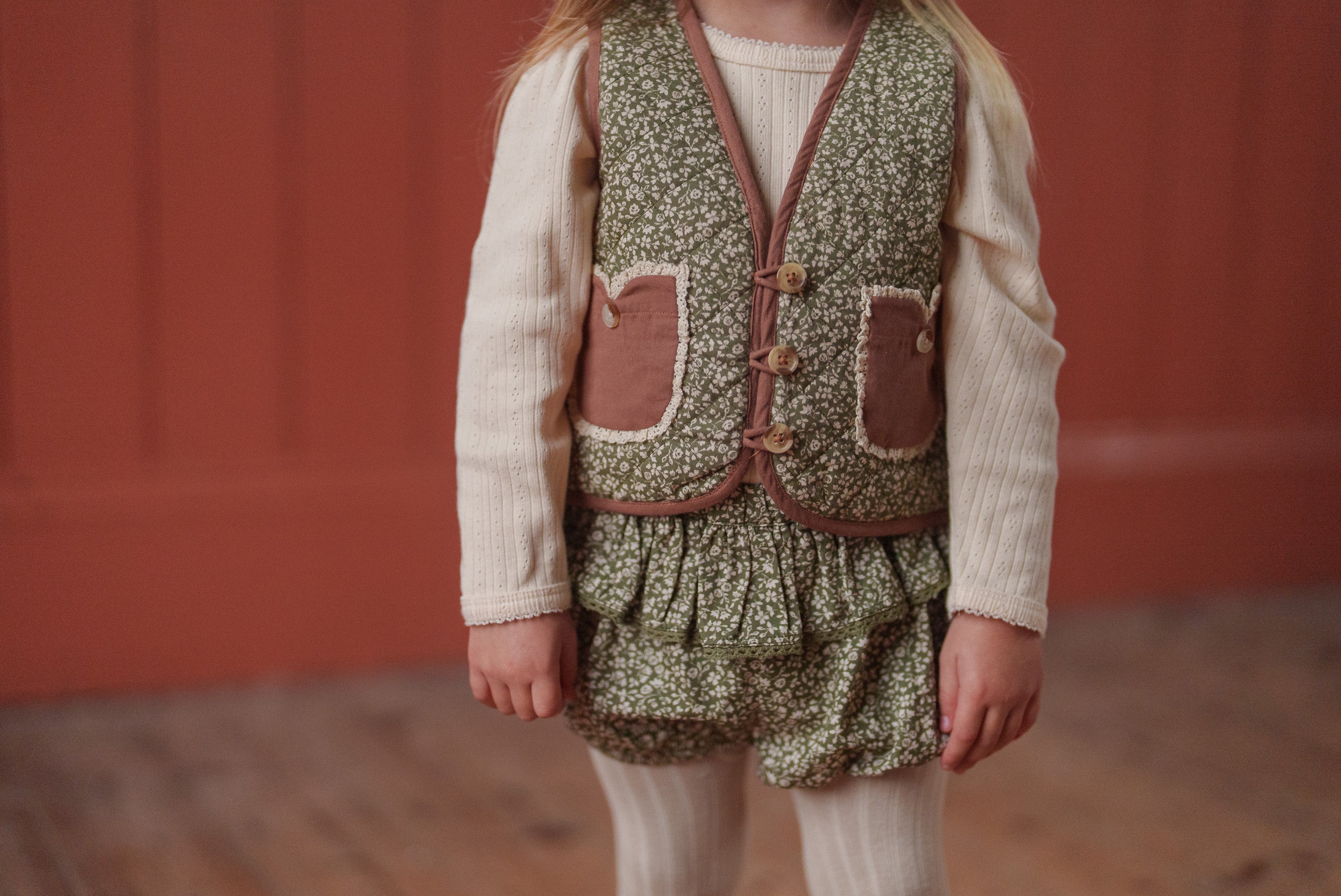 Child wearing a vintage style green floral romper made from organic cotton with a cream long-sleeve top and cream tights, showcasing sustainable children's clothing.