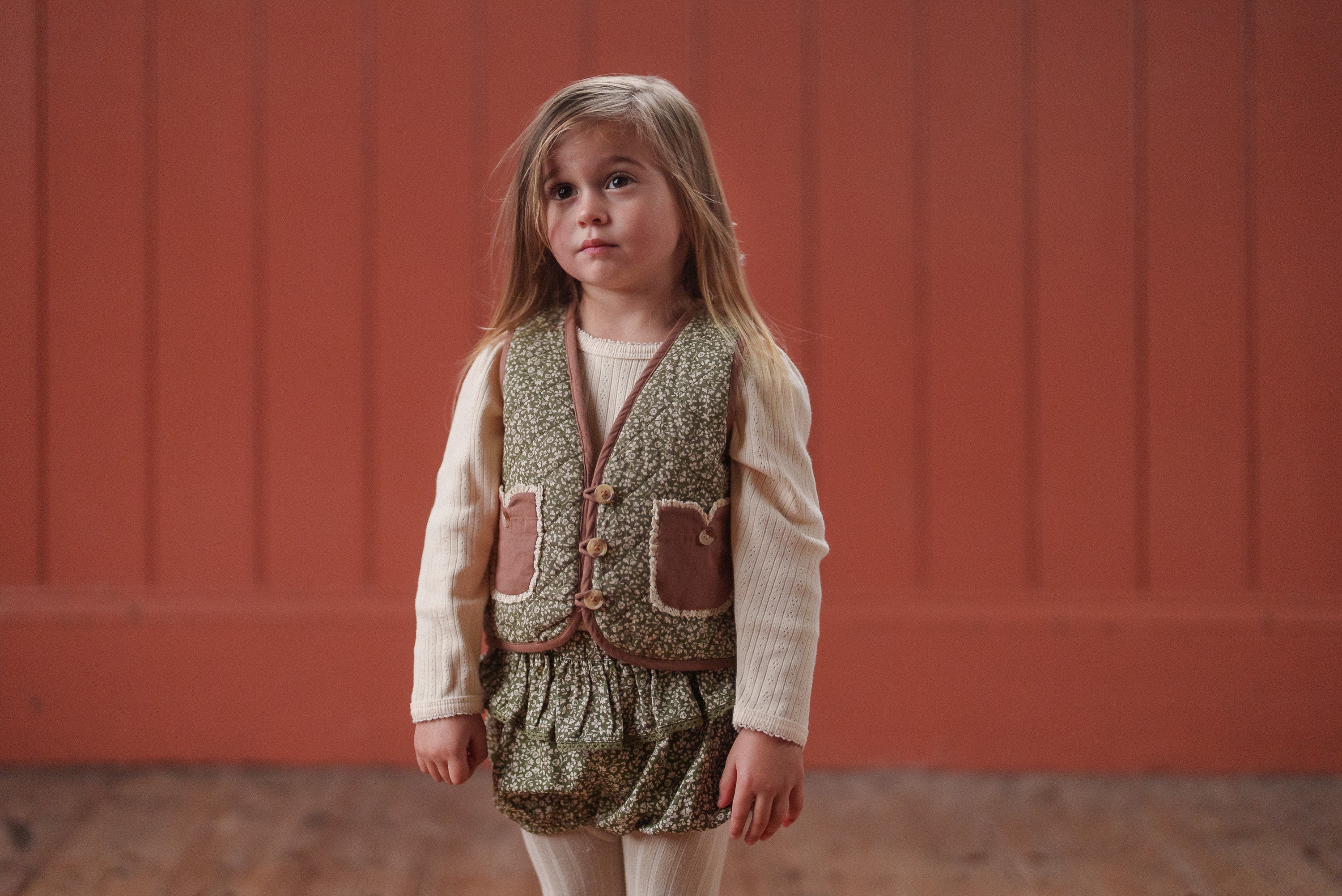 Young girl wearing vintage style children's clothing in organic cotton with a green floral romper and textured vest, showcasing ethically made sustainable kids clothing.