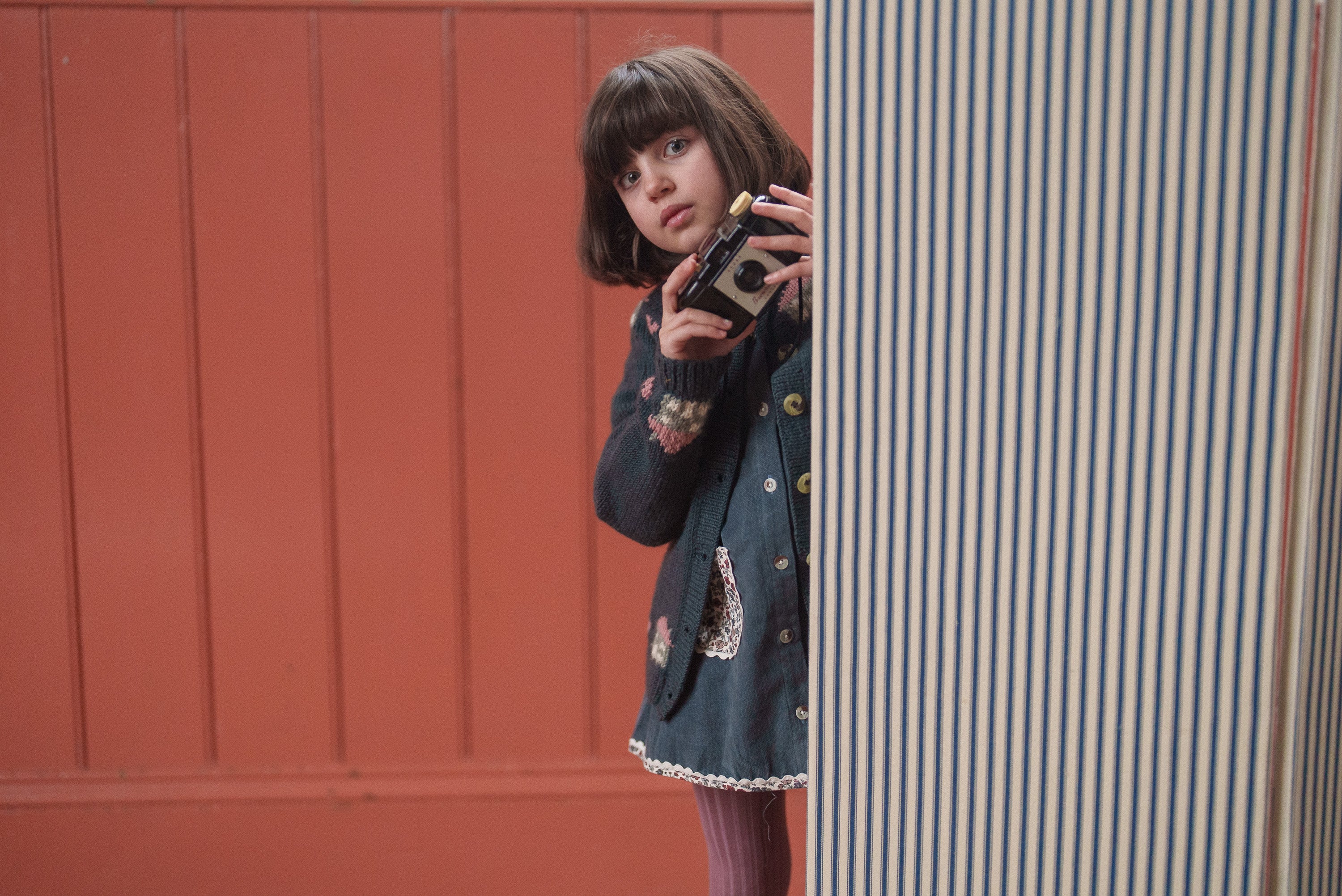 Child wearing a vintage-inspired multicolor blue merino hand-knitted cardigan, holding a camera, showcasing sustainable and organic children's clothing with a traditional style.