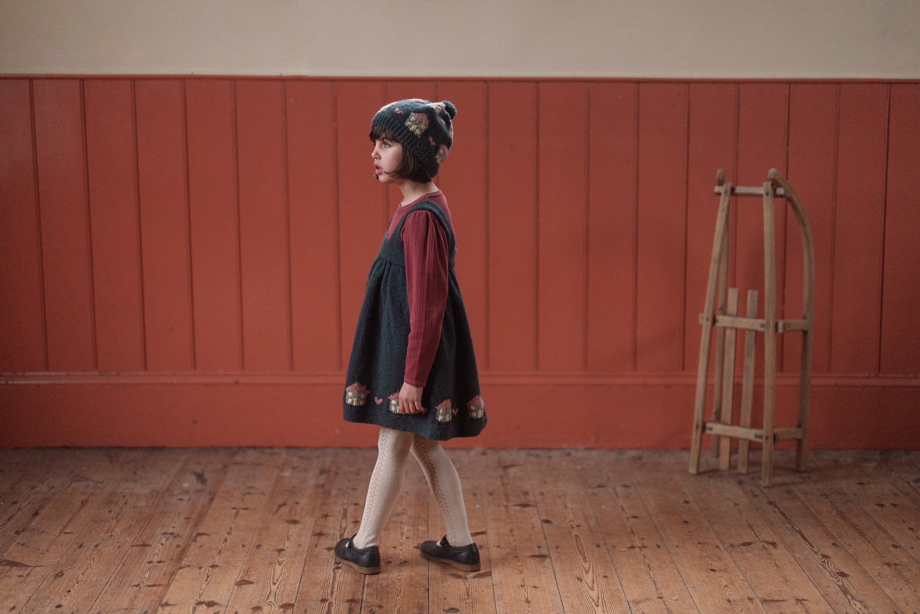 Young child wearing a hand-knitted multicolor Merino bobble hat and vintage-style dress, standing on wooden floor against a warm-toned wall in sustainable kids clothing.