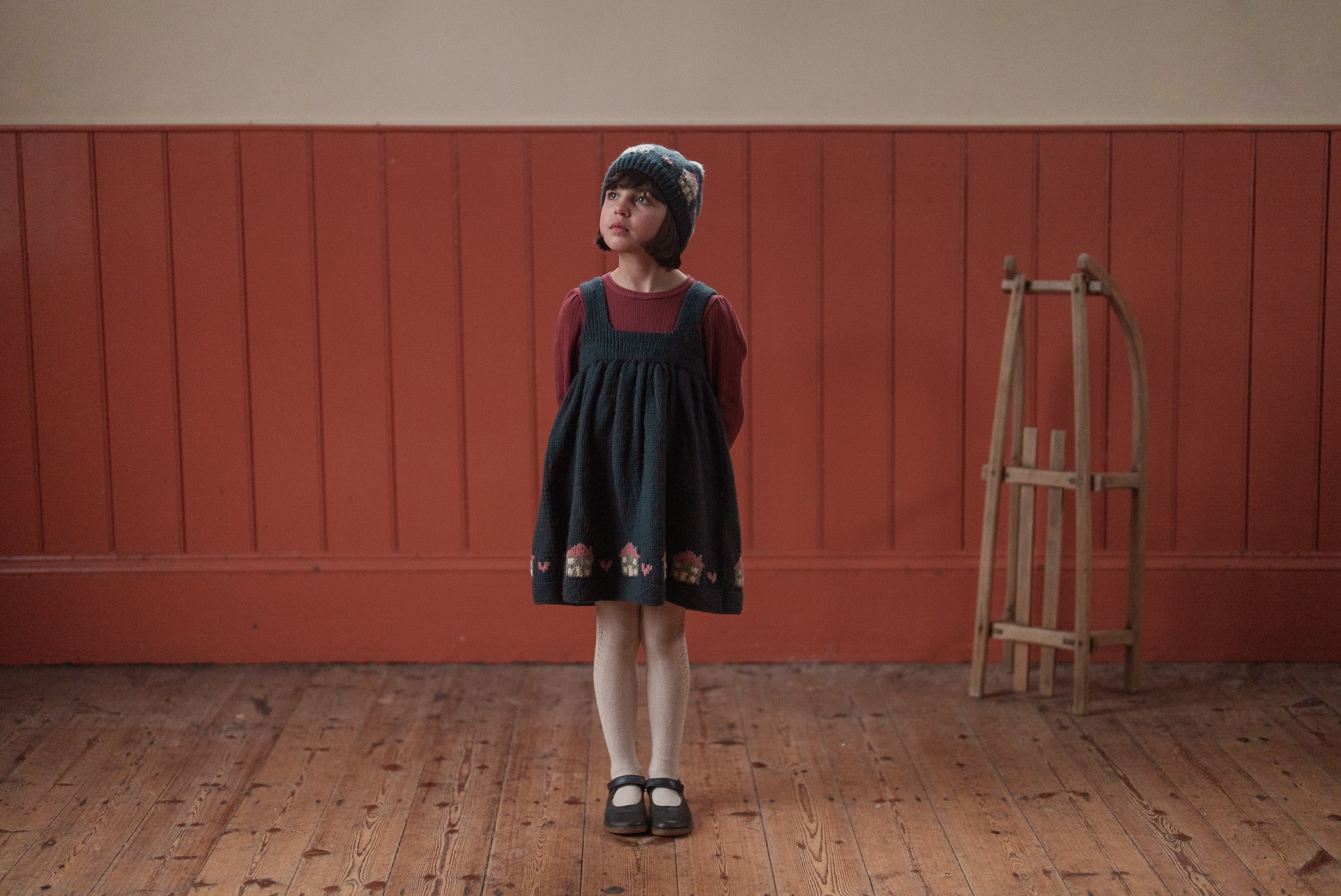 Young girl wearing a vintage-style navy merino pinafore dress with floral embroidery, paired with a burgundy long-sleeve top and a matching knitted hat, standing on a wooden floor indoors.