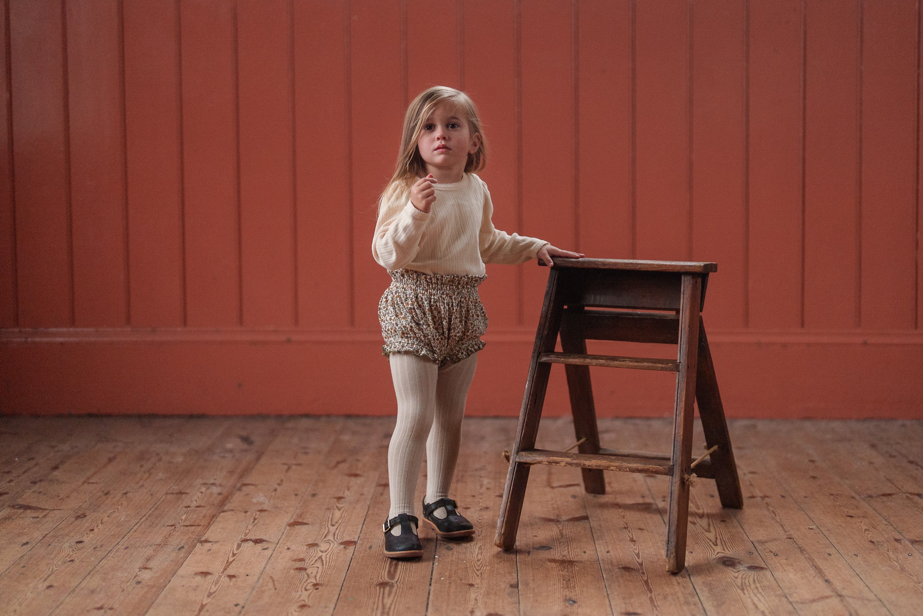 Young child wearing a cream organic cotton pointelle T-shirt and vintage-style bloomers, standing by a wooden step ladder on a wooden floor with a warm orange background.