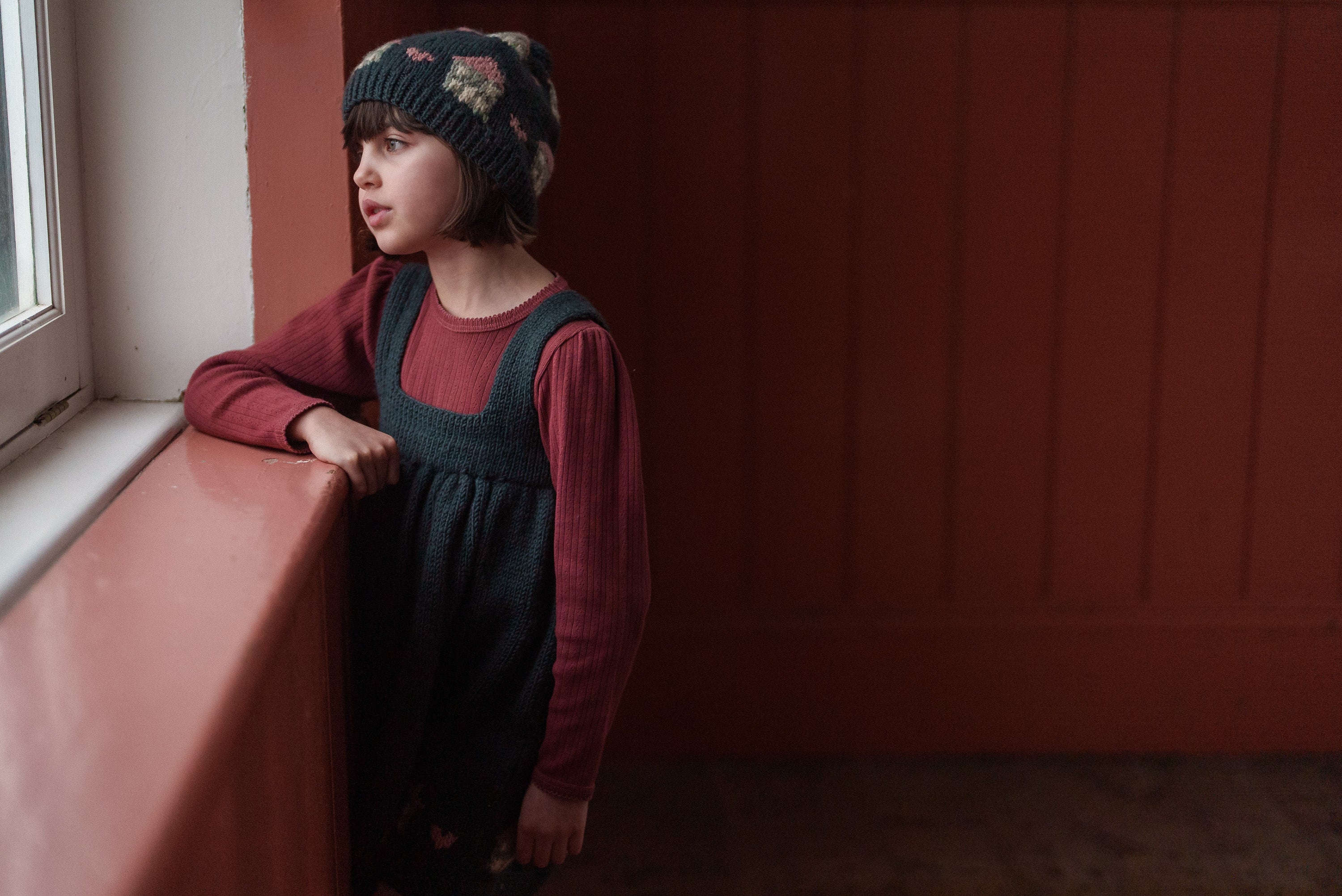 Young girl wearing a red organic cotton pointelle T-shirt layered under a dark vintage-style dress and a knitted hat, looking thoughtfully out a window in a warm, natural setting.