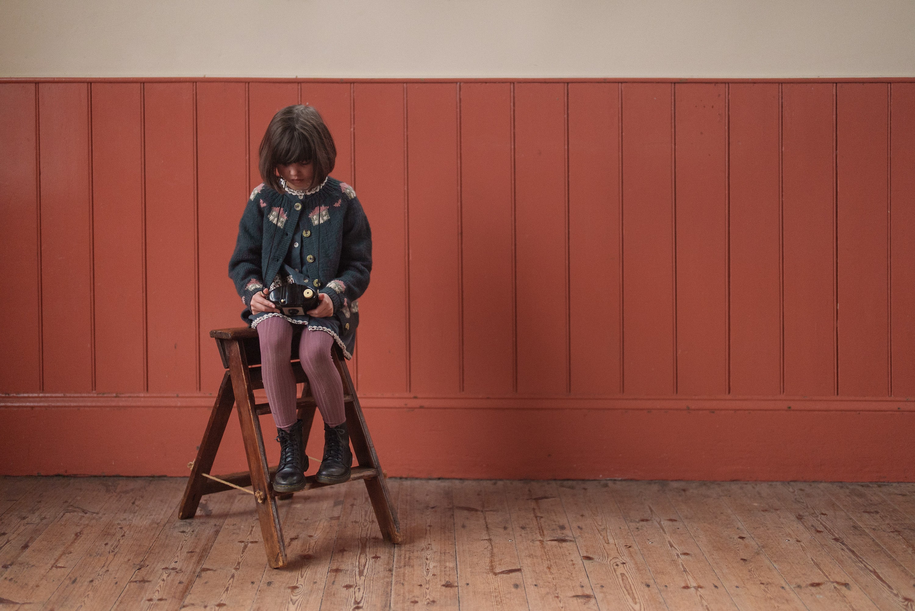 Child wearing a multicolor blue hand-knitted merino cardigan sitting on a wooden folding chair in a vintage-style room with natural lighting, showcasing sustainable kids clothing.