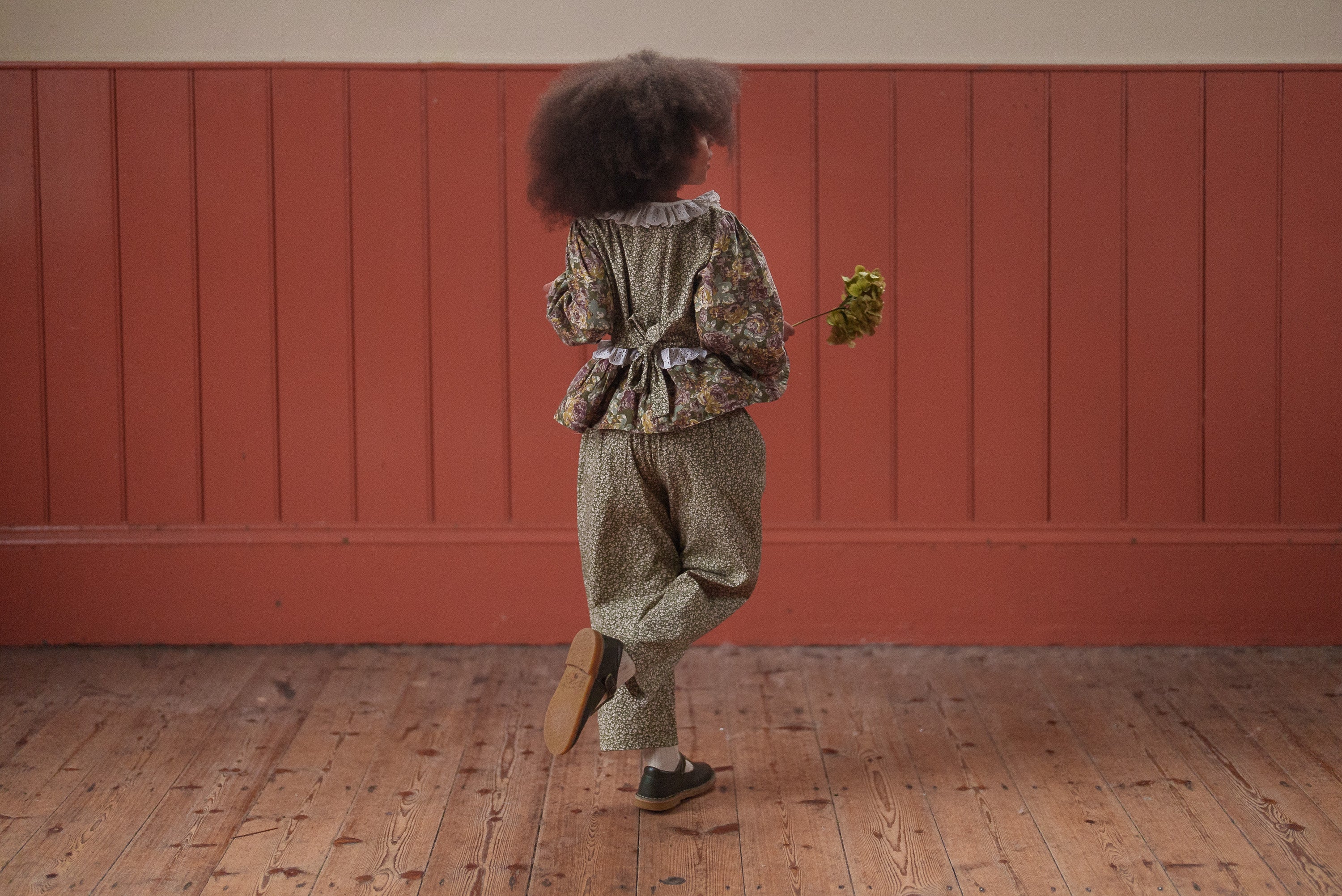 Child wearing a vintage style organic cotton blouse and trousers with a floral pattern in green and pink, walking indoors on a wooden floor holding a small bunch of flowers.