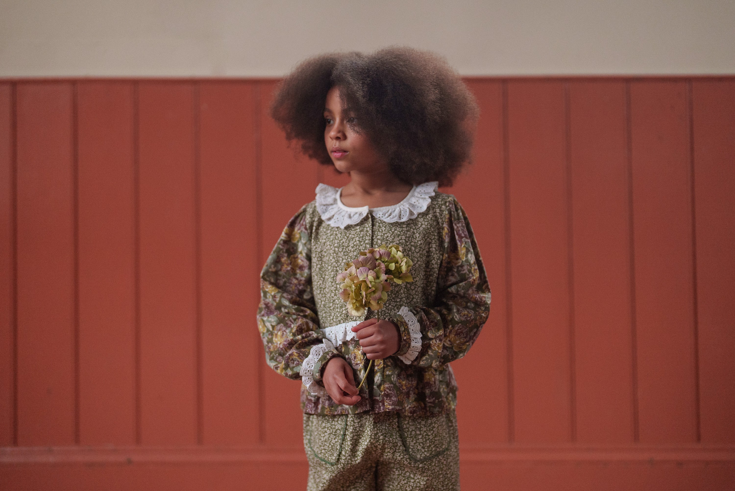 Young girl wearing a vintage style green and pink floral organic cotton blouse with lace collar and cuffs, holding dried flowers in a softly lit room.