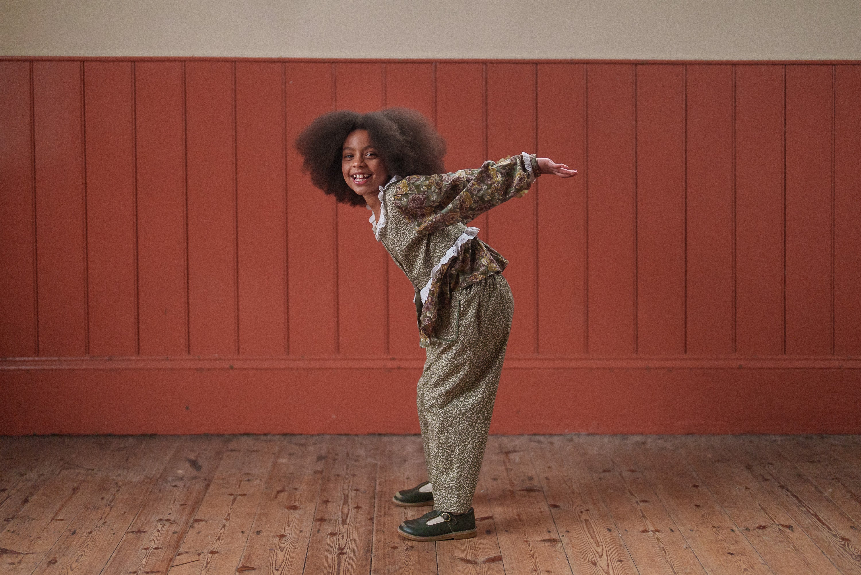 Child wearing a green and pink floral organic cotton blouse and matching trousers, smiling and playfully bending forward in a vintage style setting.