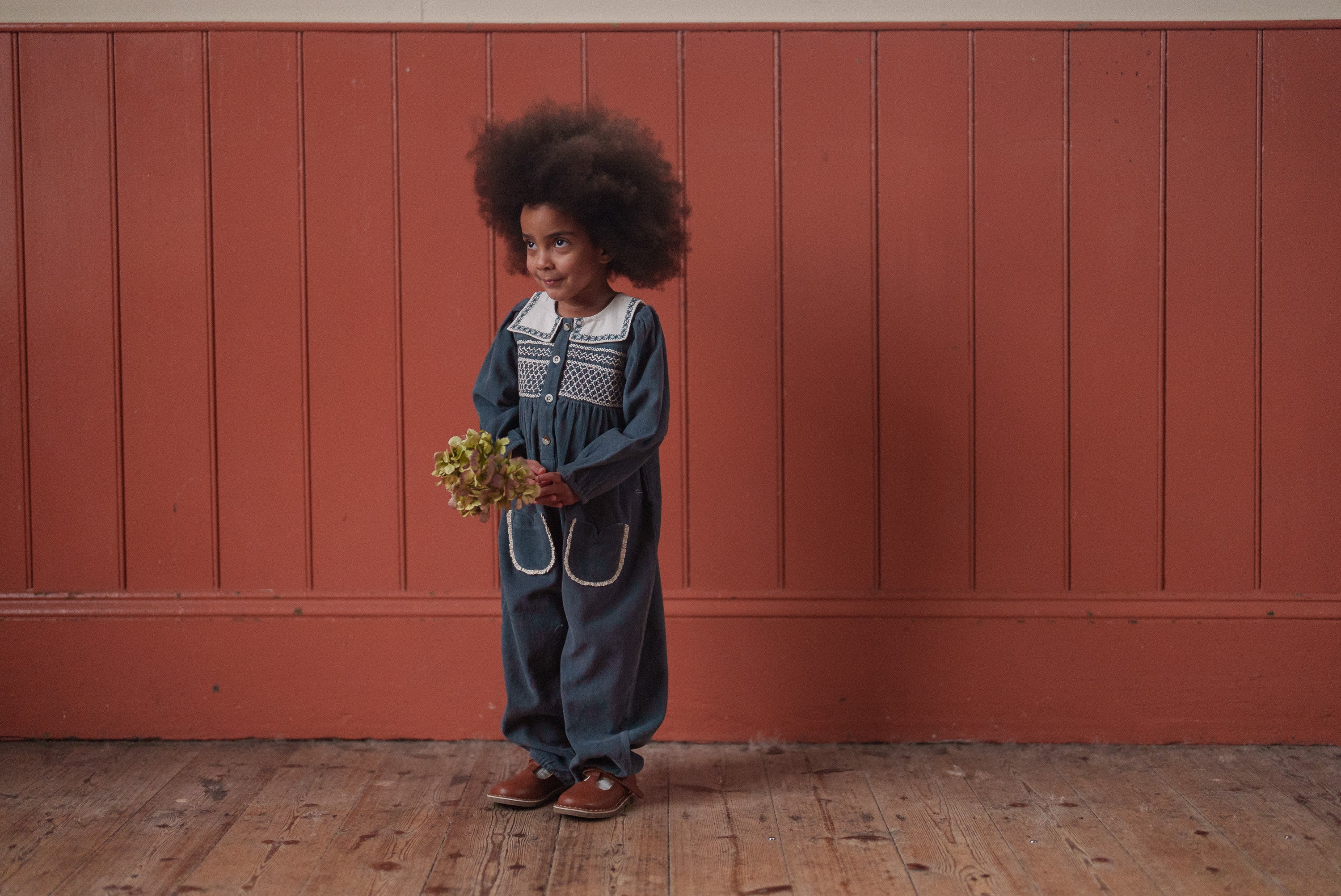 Young child wearing vintage-style organic cotton romper in deep green, holding a small bouquet, standing on a wooden floor against a warm-toned paneled wall.