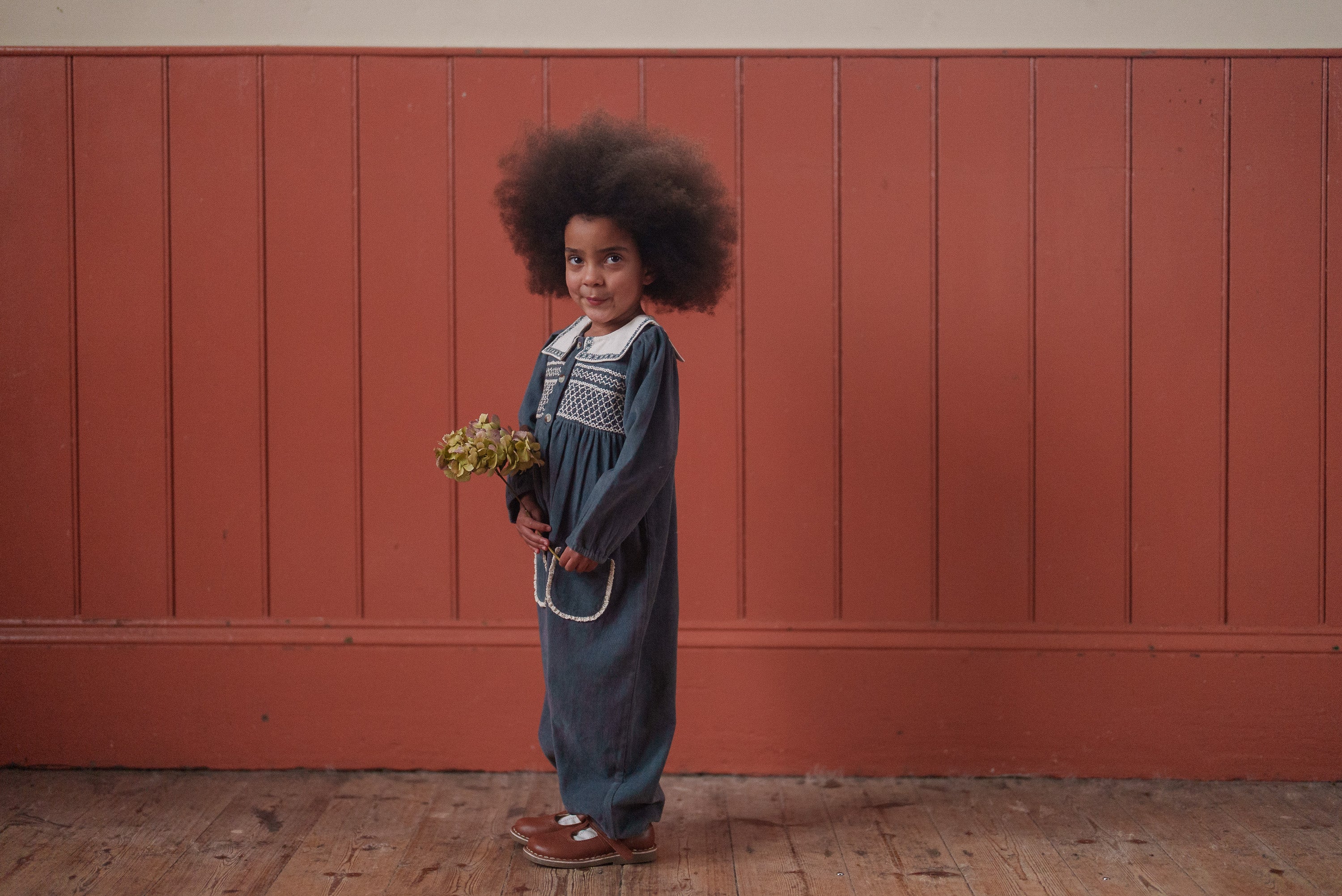 Young child wearing a vintage-style organic cotton romper in juniper holding a small bouquet, standing in front of a rustic wooden wall in natural light.