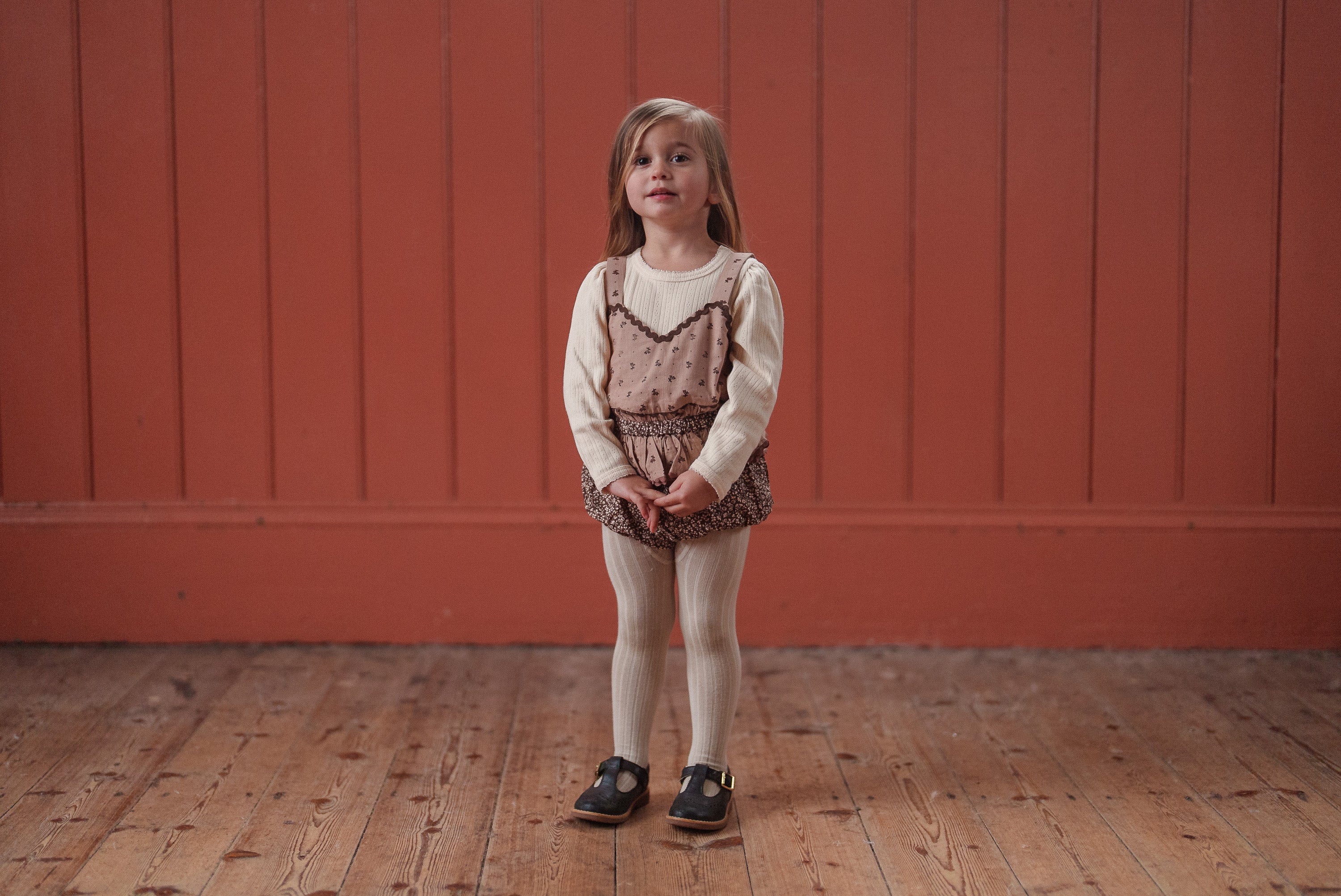 A young girl wearing a vintage-inspired brown and floral romper made from organic cotton, paired with cream tights and a cream long-sleeve top, standing on a wooden floor against a terracotta wall.