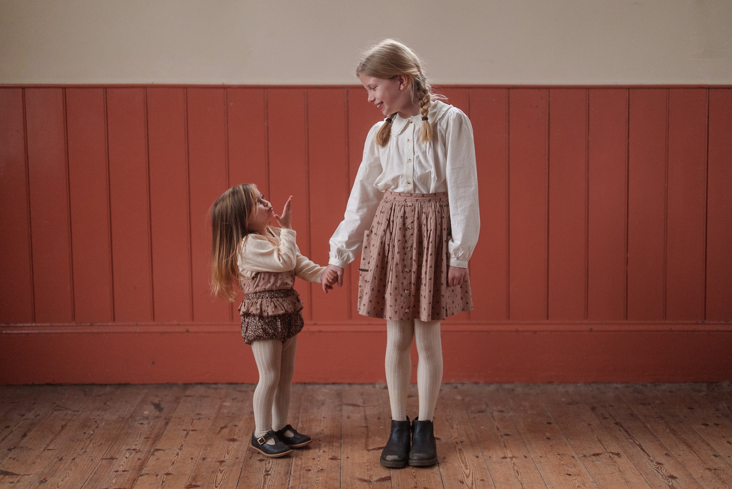 Two young girls holding hands indoors, wearing vintage style children's clothing made from organic cotton in brown and floral patterns, reflecting natural and ethical fashion.