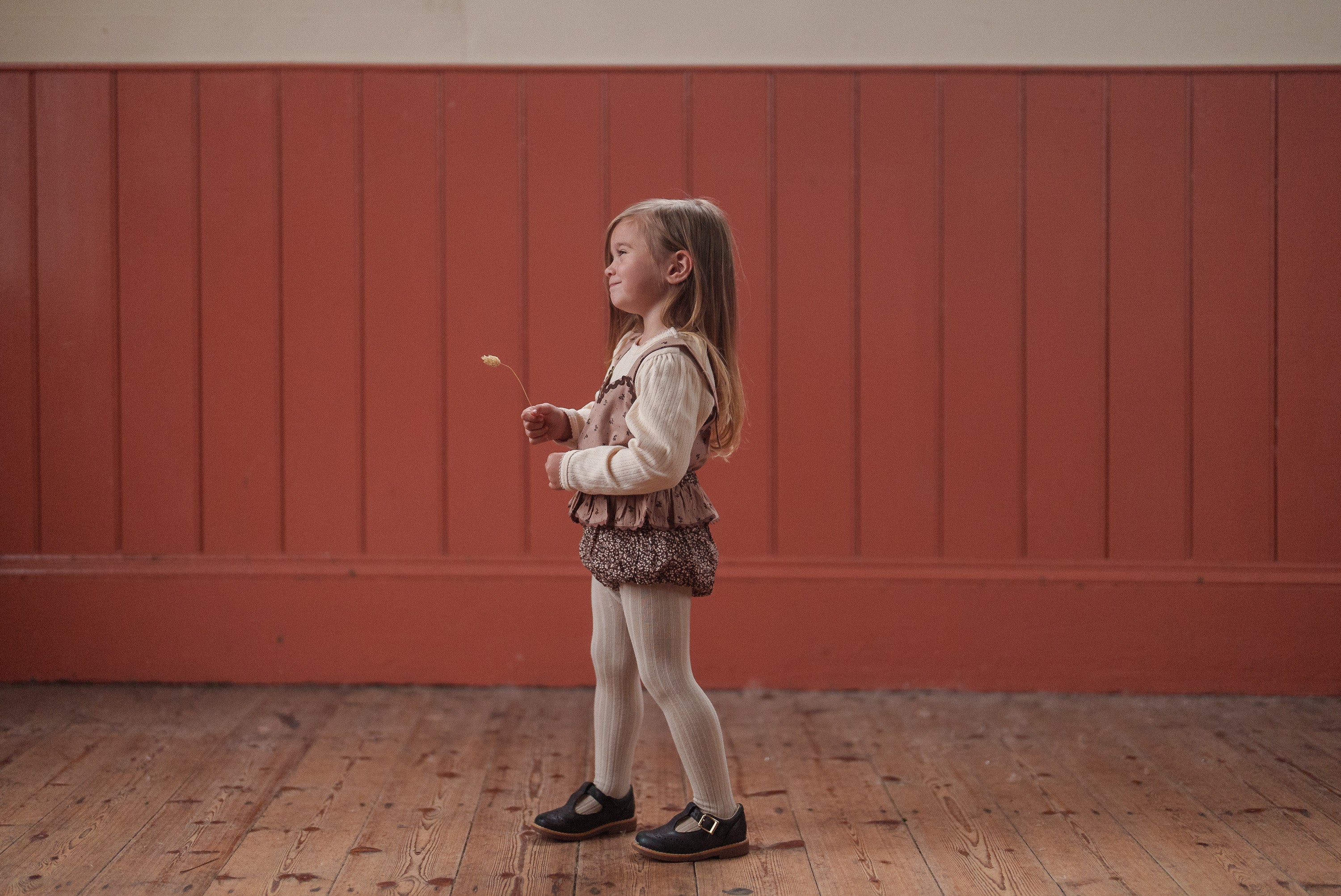 Young girl wearing a vintage-style brown floral romper made from organic cotton, standing on a wooden floor against a warm red wall in natural light.