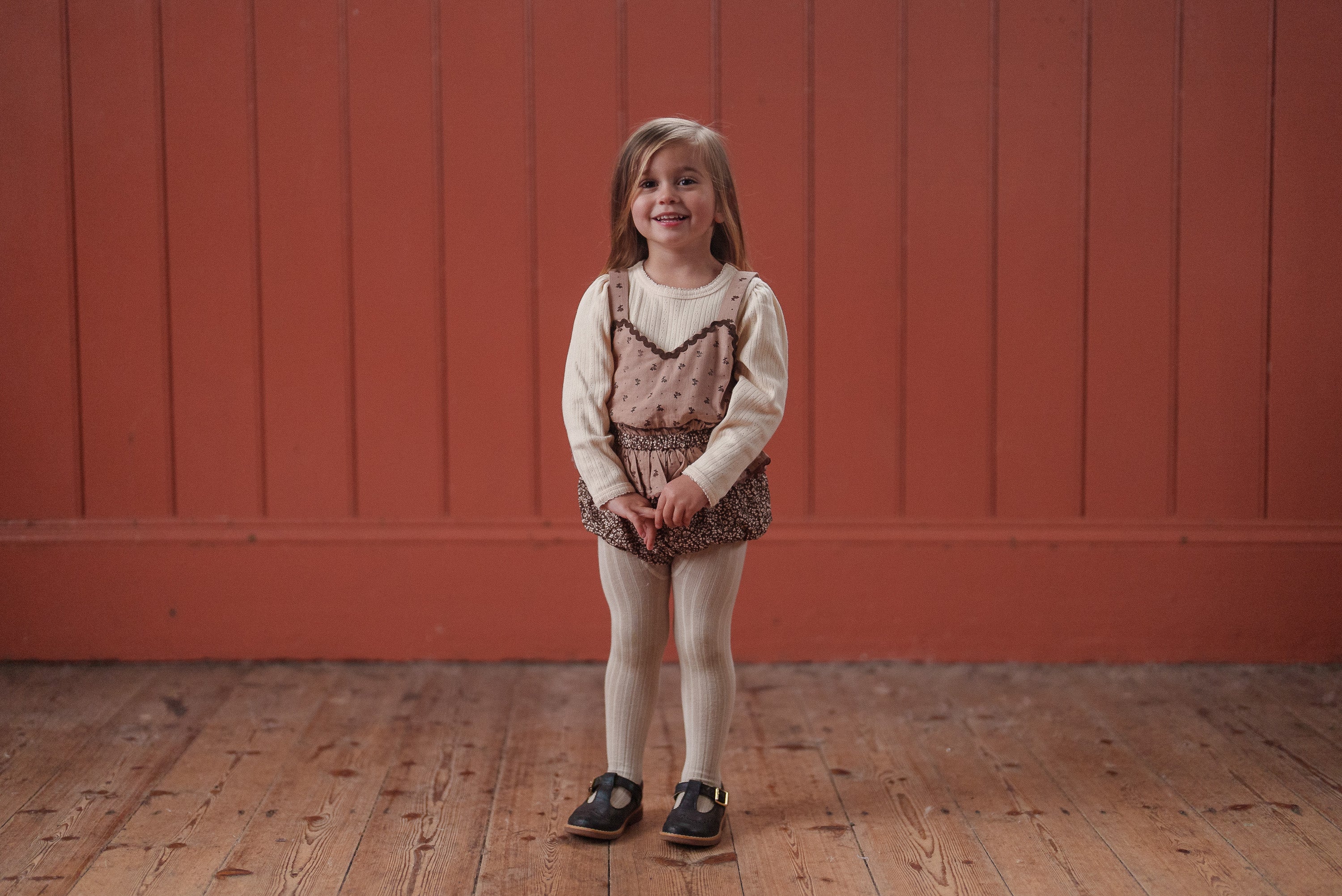 Smiling young child wearing a vintage style pink and brown floral romper made from organic cotton, paired with cream tights and black shoes, standing on wooden floor.