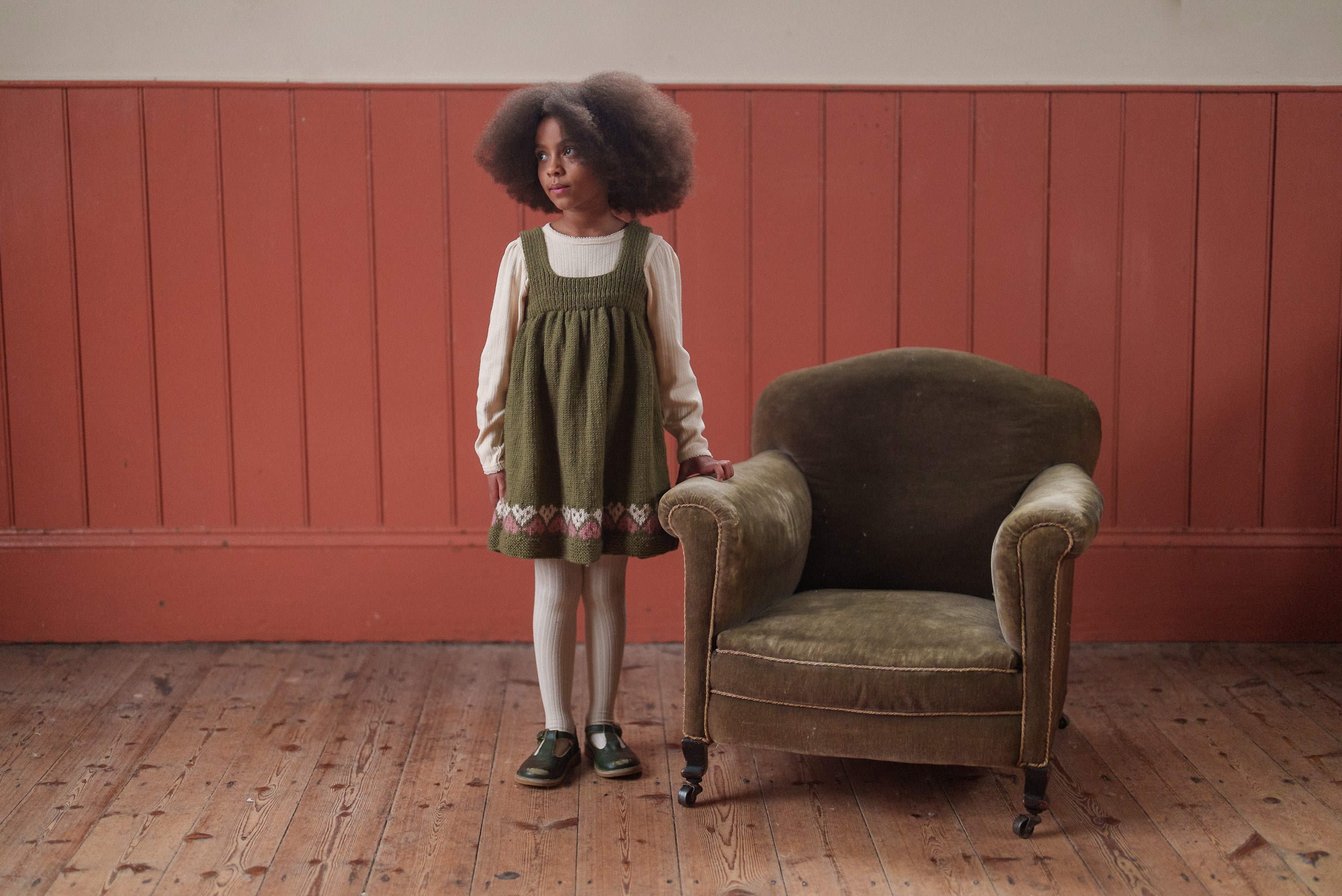 Young girl wearing a vintage-inspired hand-knitted pinafore dress in earthy green with heart details, standing next to an olive green armchair on wooden floor against a warm terracotta wall.