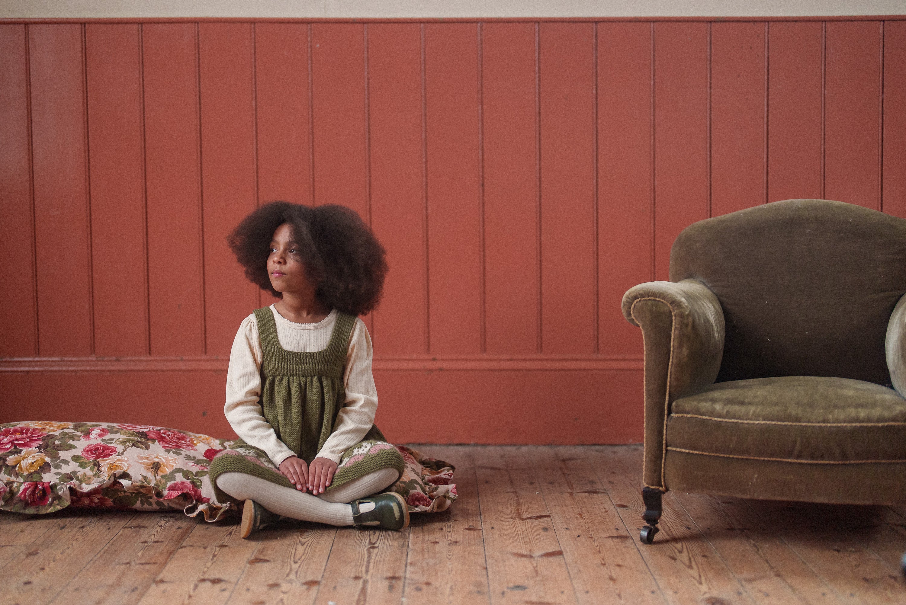 Young girl wearing a vintage-style hand-knitted pinafore dress in natural merino wool, sitting on a floral cushion against a warm-toned wooden wall, next to a classic armchair