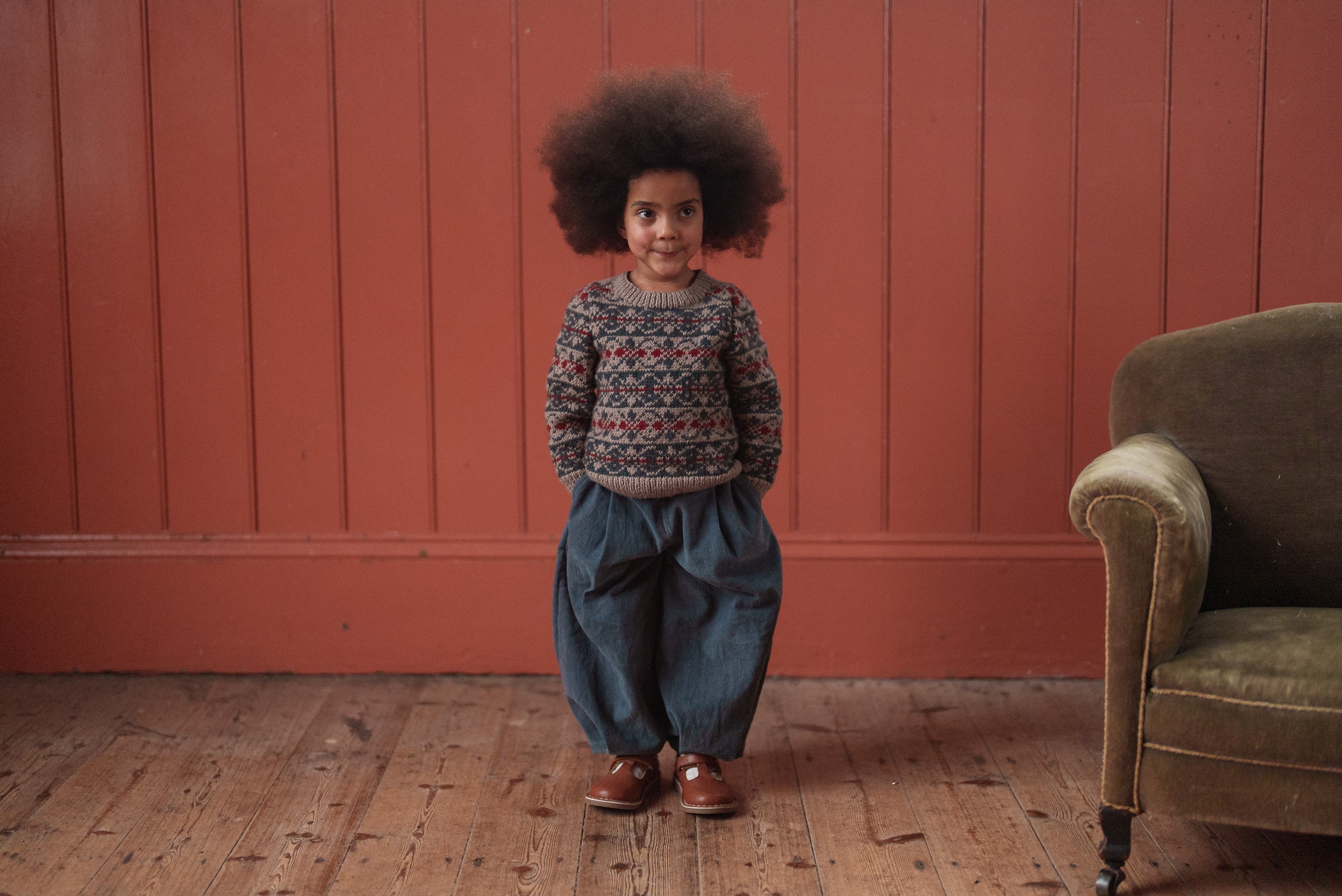 Child wearing a multicolor hand-knitted jumper made from soft merino wool, paired with loose vintage-style navy pants in a cozy, natural setting.