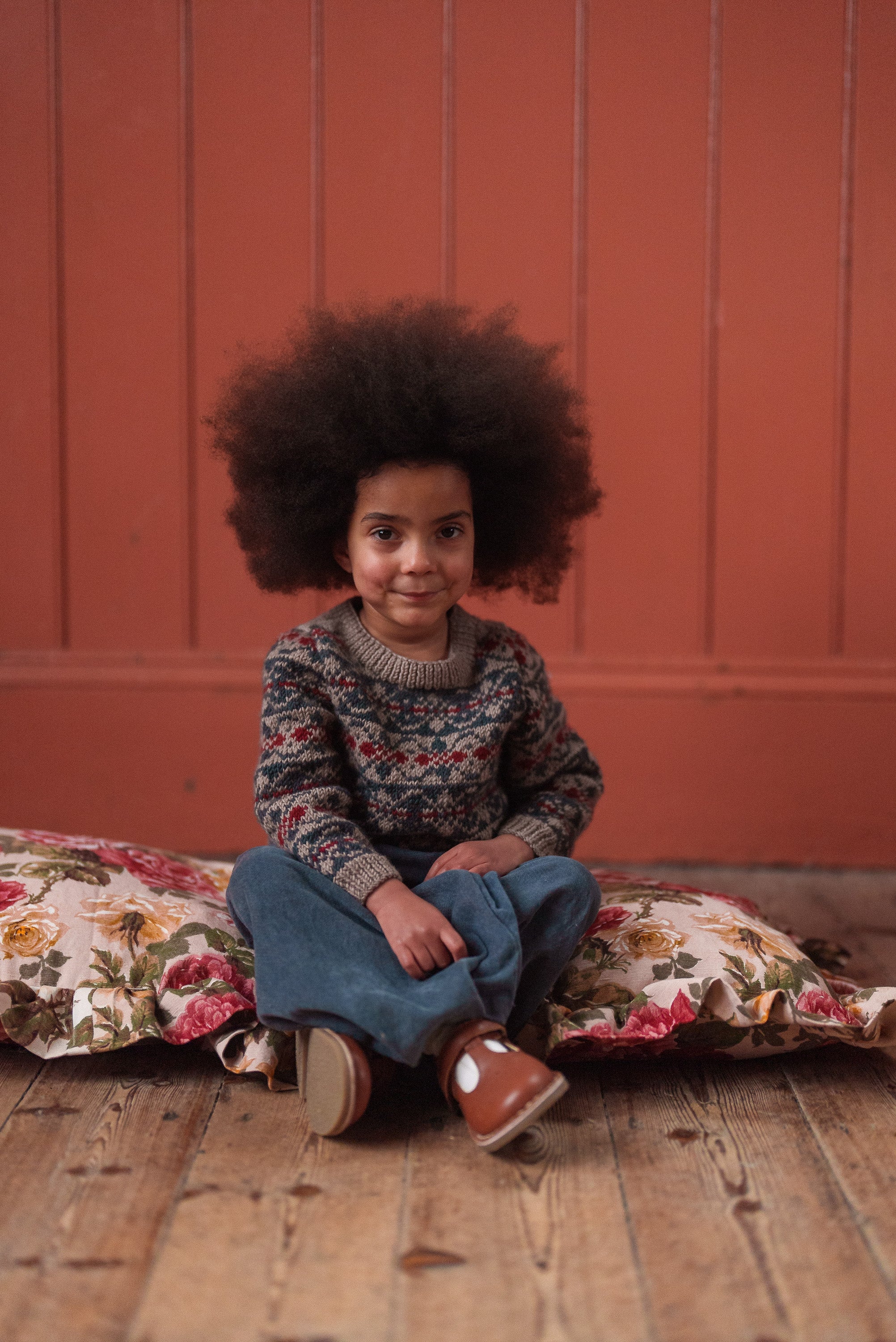 Child wearing a multicolor hand-knitted Fairisle jumper made from soft Merino wool, sitting on floral cushions, showcasing sustainable vintage style kids clothing.