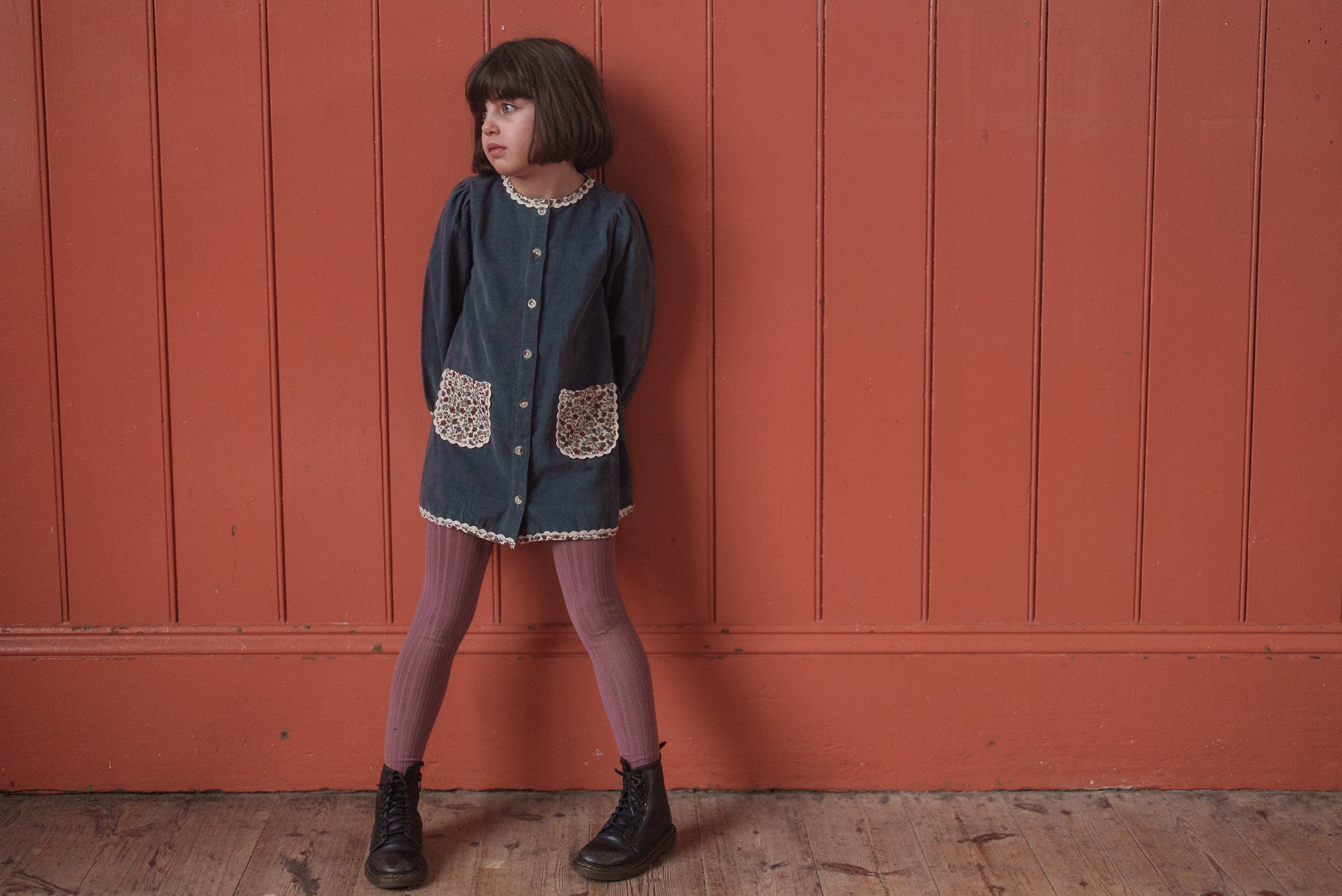 Young girl wearing a vintage-style dark blue velvet dress with floral trim and pockets, standing against a wooden wall, showcasing organic and sustainable children's clothing.