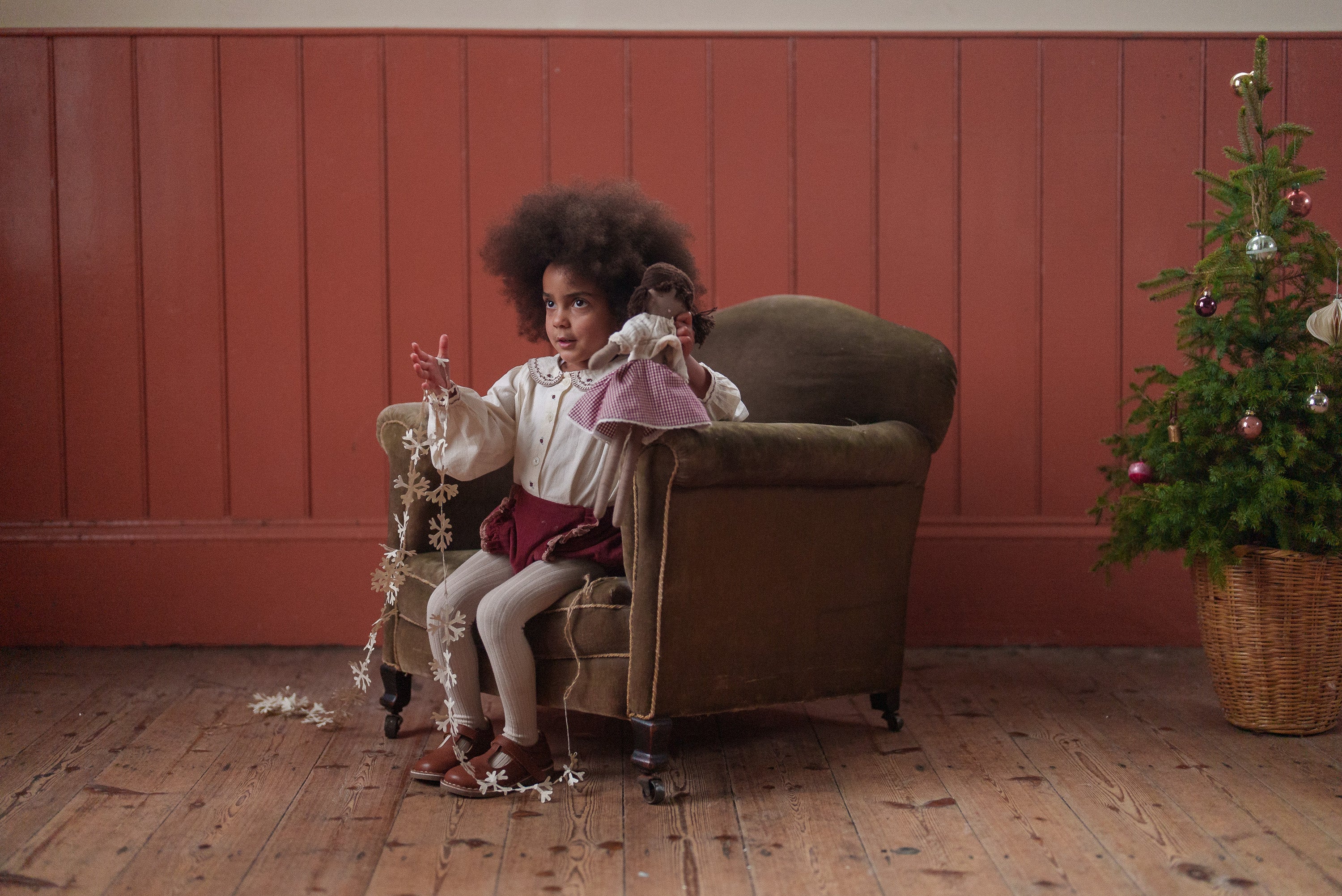 Child wearing sustainable vintage style clothing in pink velvet bloomers, sitting on a green armchair with a doll and decorative garland by a Christmas tree.