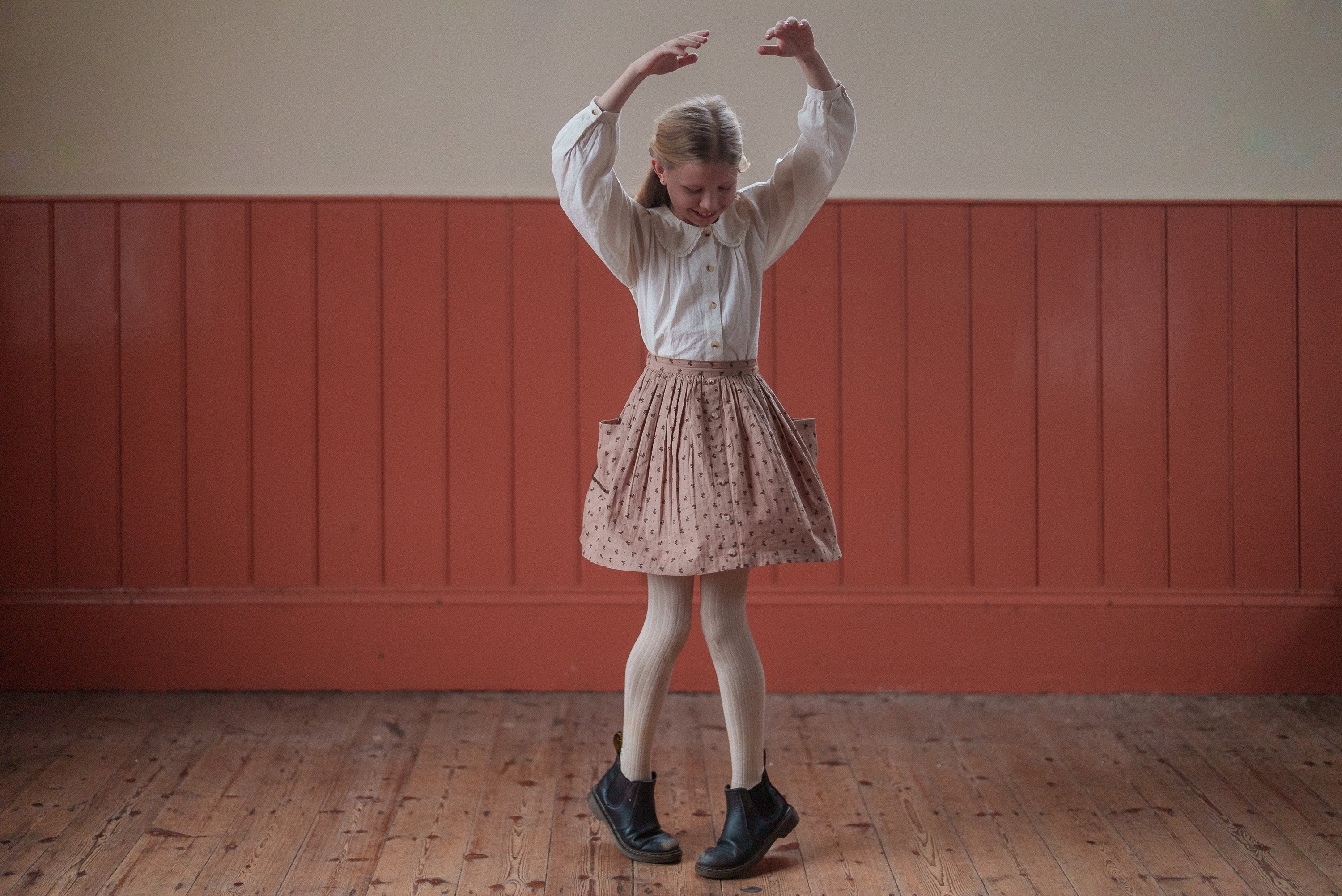 Young girl wearing a pink floral organic cotton skirt and white blouse, dancing joyfully in a vintage-style room, showcasing sustainable and ethically made children's clothing.