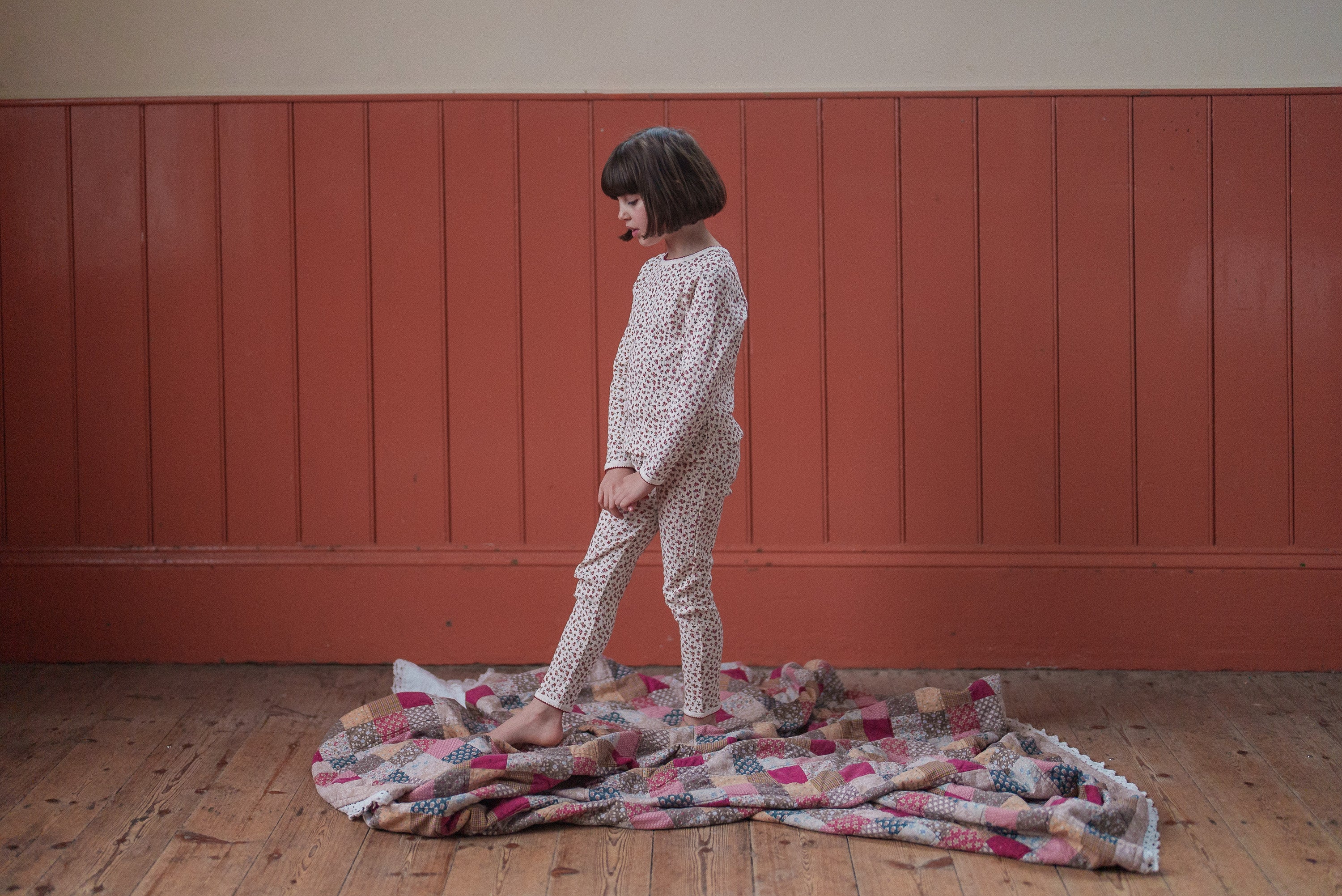 Child wearing soft organic jersey cotton pyjamas with a delicate floral pattern, standing barefoot on a vintage patchwork blanket in a cozy wooden room.