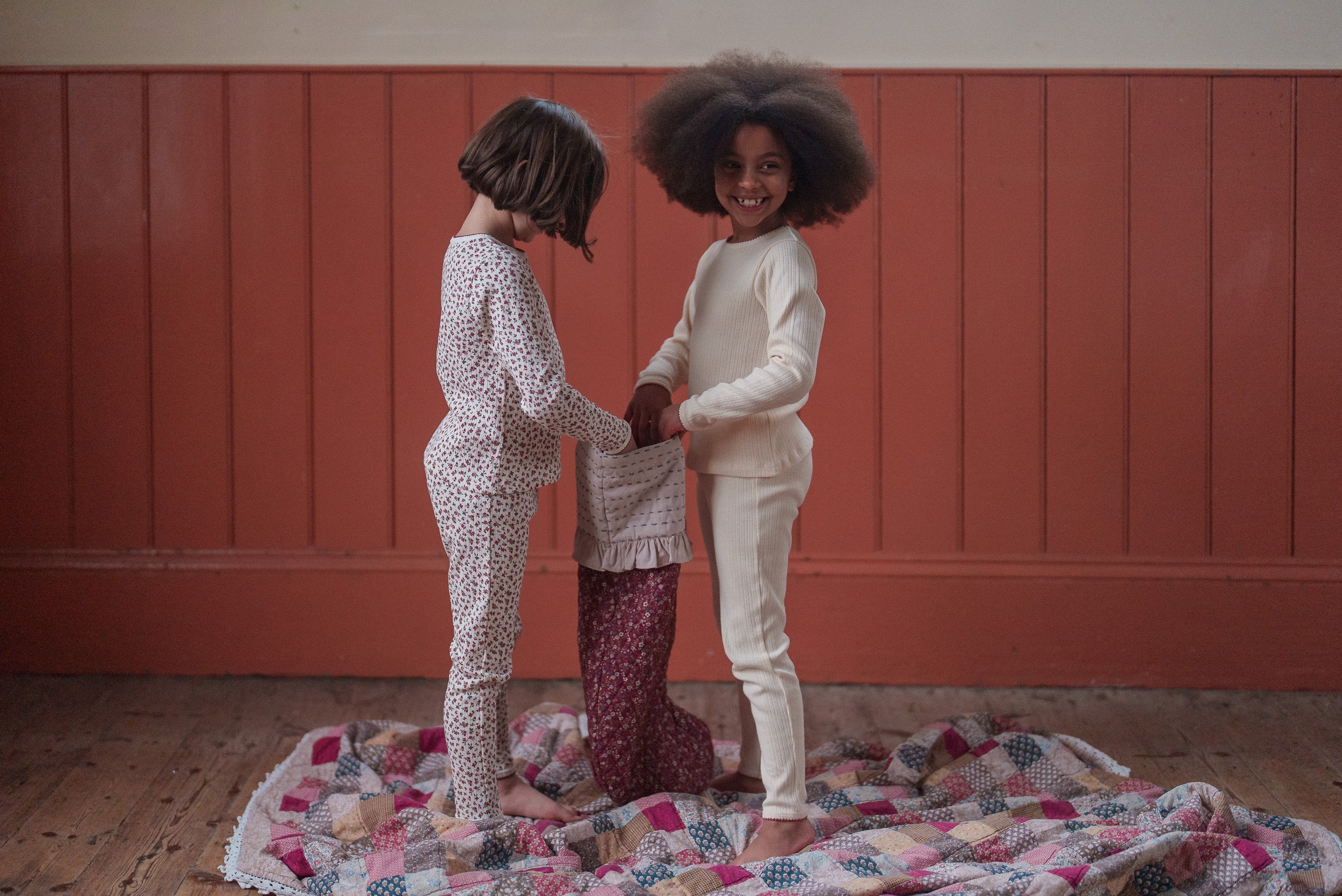 Two children wearing soft, natural cotton pajamas in warm neutral tones, standing on a patchwork quilt in a cozy vintage-style room, embodying sustainable and ethical kids clothing.