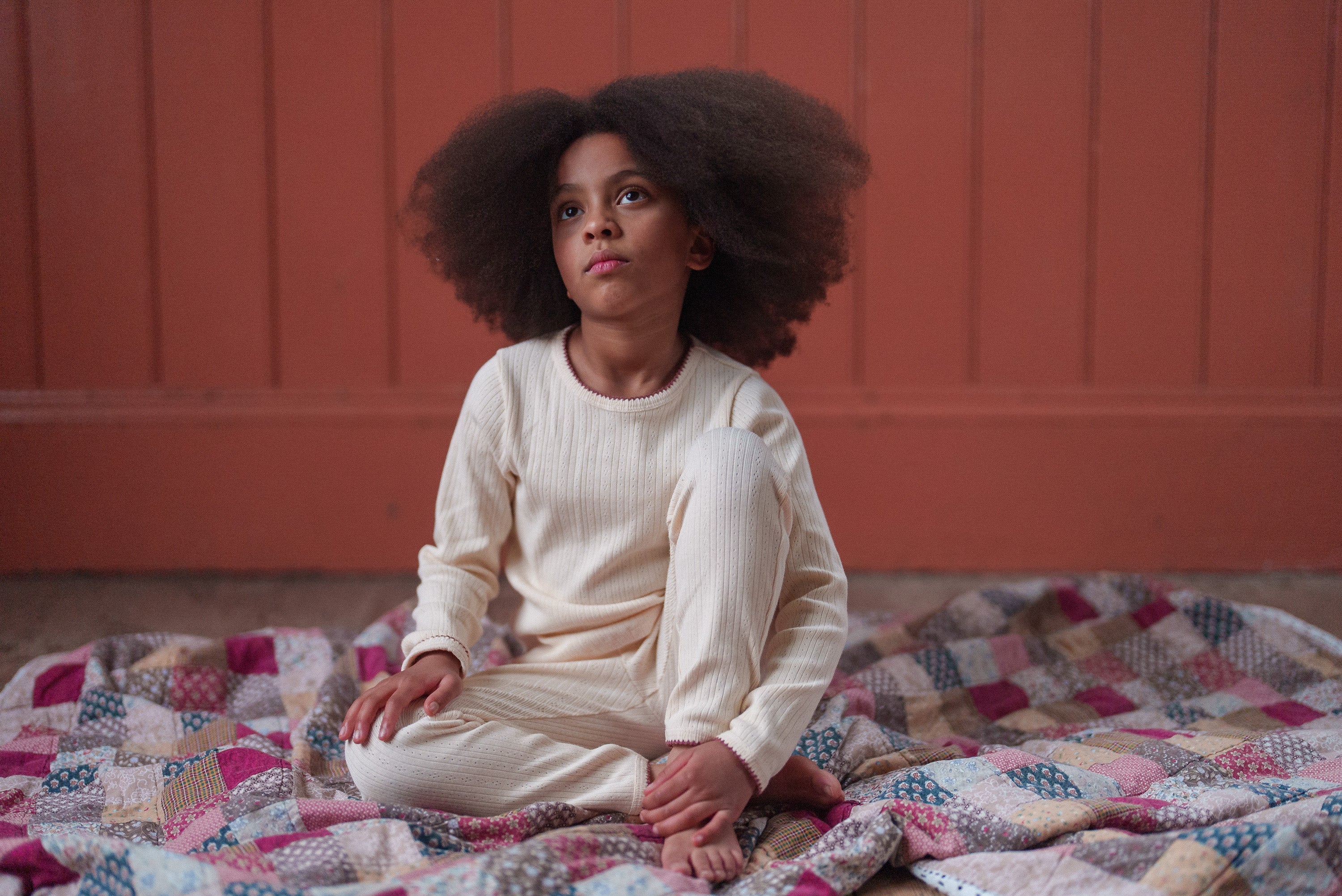 Young girl sitting on a vintage patchwork quilt wearing soft buttermilk-colored organic cotton pyjamas in a natural, timeless style.