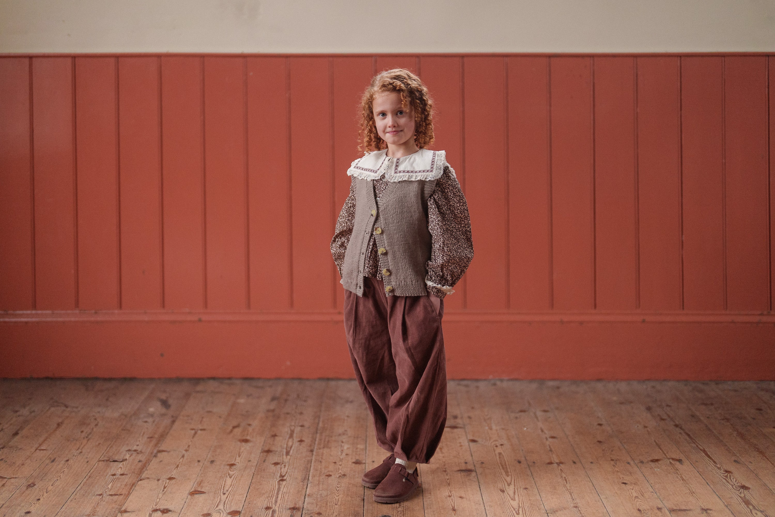 A young child wearing vintage style children's clothing including a multicolor beige and brown hand-knitted wool vest over a patterned blouse with a white lace collar, standing on a wooden floor in a warm, natural setting.