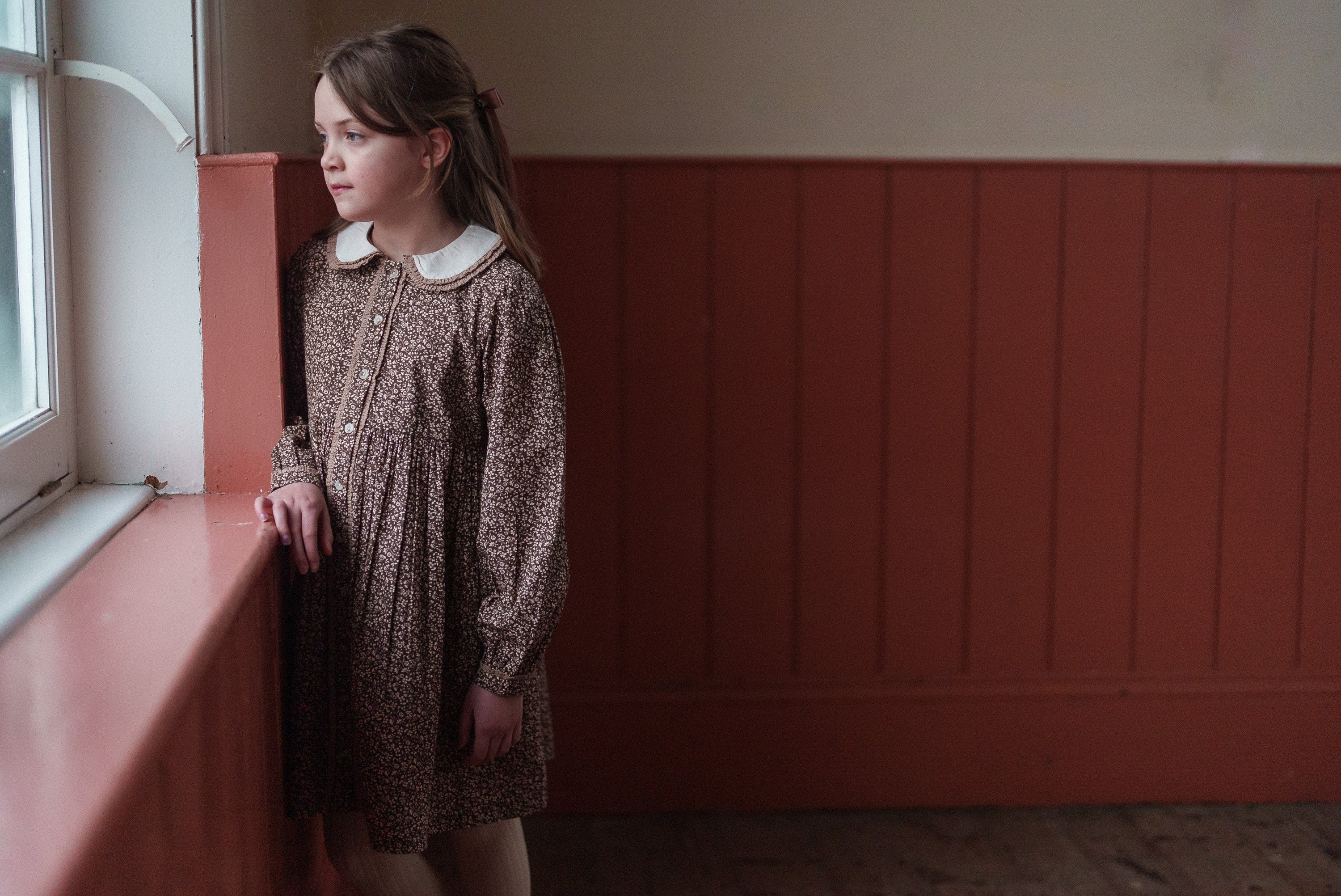 Young girl in a vintage-style brown and cream floral organic cotton dress standing by a window in a softly lit room, showcasing sustainable children's clothing.