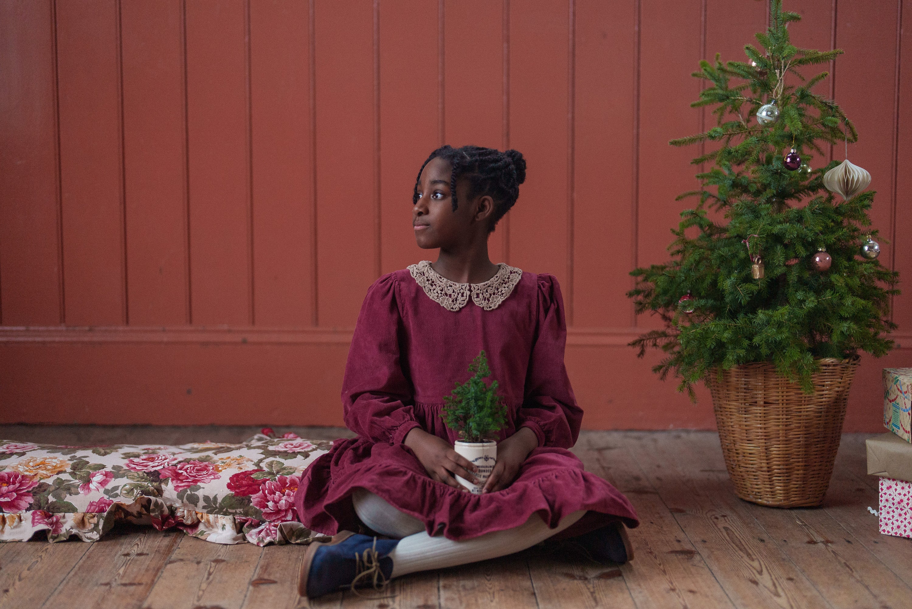 Young girl wearing a red velvet vintage-style dress with a lace collar, sitting on a wooden floor holding a small potted plant near a decorated Christmas tree in a cozy, natural setting.