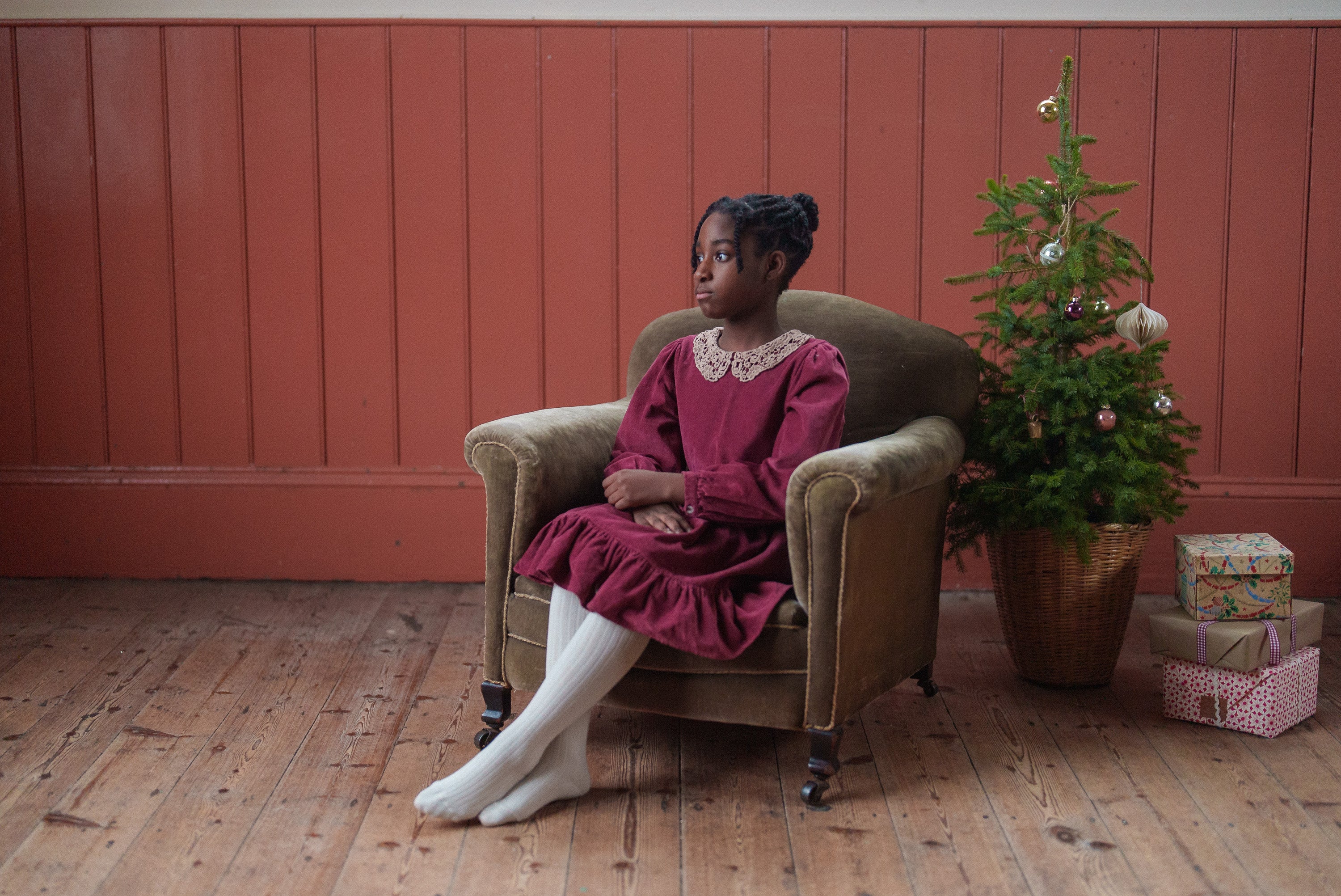 Young girl wearing a red velvet dress made from organic cotton, sitting on a vintage-style armchair next to a small Christmas tree with wrapped gifts, showcasing sustainable kids clothing.