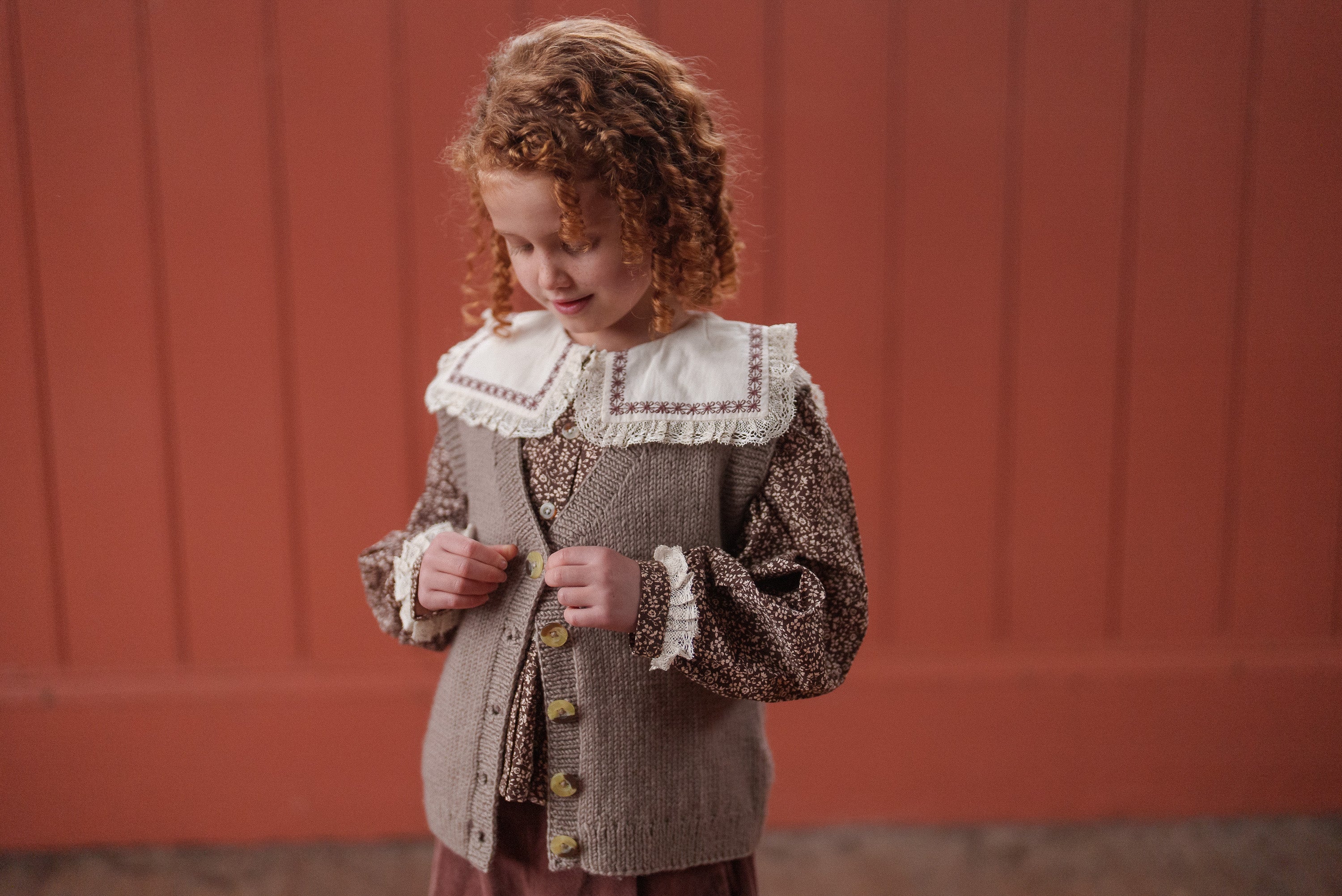 Young girl wearing a vintage style floral blouse in brown, beige, and cream with a knitted vest, showcasing sustainable, organic cotton children’s clothing designed with traditional details.