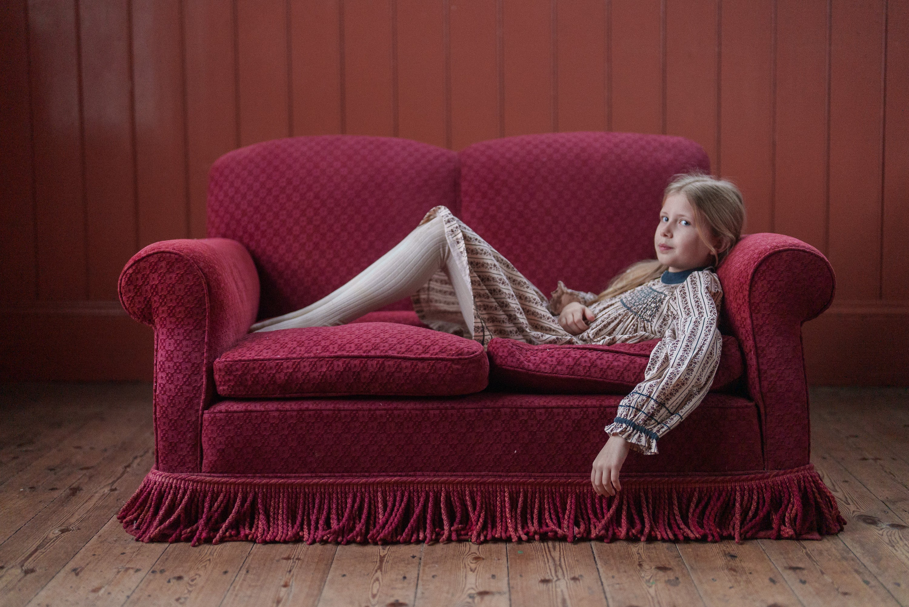 Young girl wearing a vintage style organic cotton dress reclining on a deep red vintage sofa in a rustic wooden room, showcasing sustainable children's clothing.