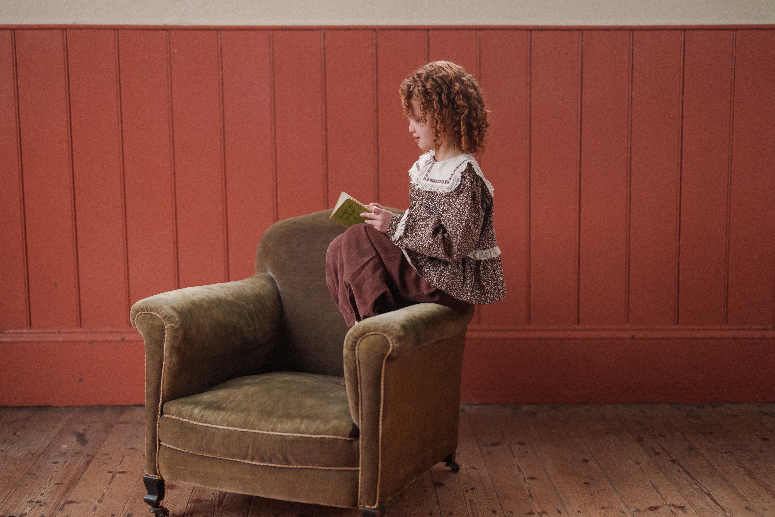 Young girl wearing a vintage-style brown floral blouse made from organic cotton, sitting on an olive green armchair against a rustic wooden wall.