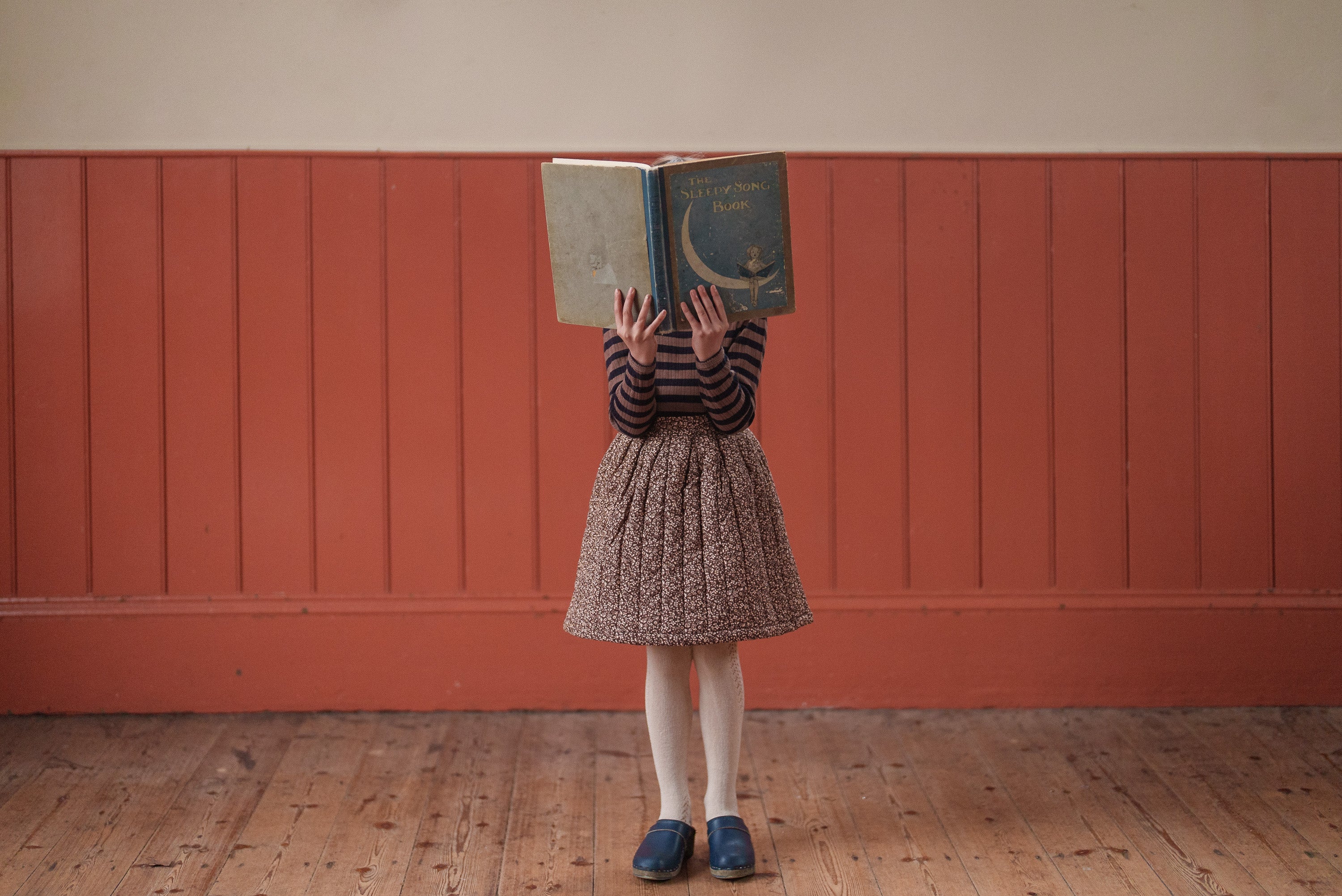 Child wearing slim fit navy and beige striped organic cotton T-shirt and vintage style skirt, holding an open book covering their face in a warm, natural setting