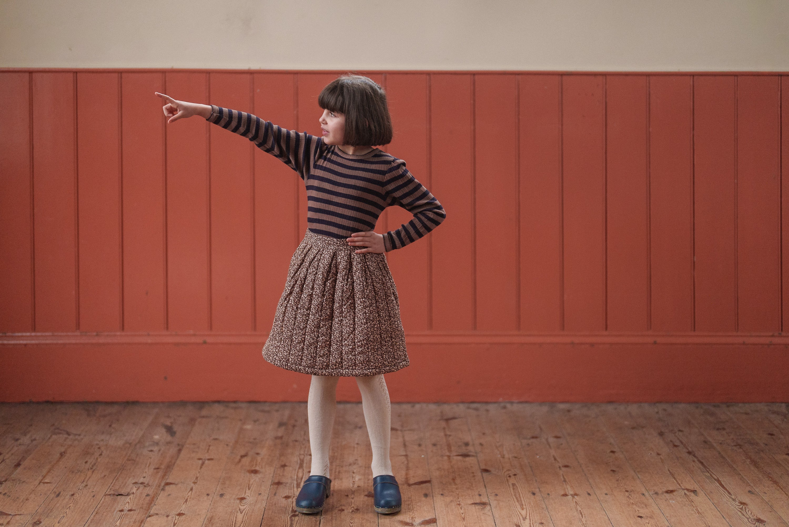 Child wearing a vintage-style striped organic cotton T-shirt and textured skirt, standing on a wooden floor against a warm-toned wall, showcasing sustainable children's clothing.