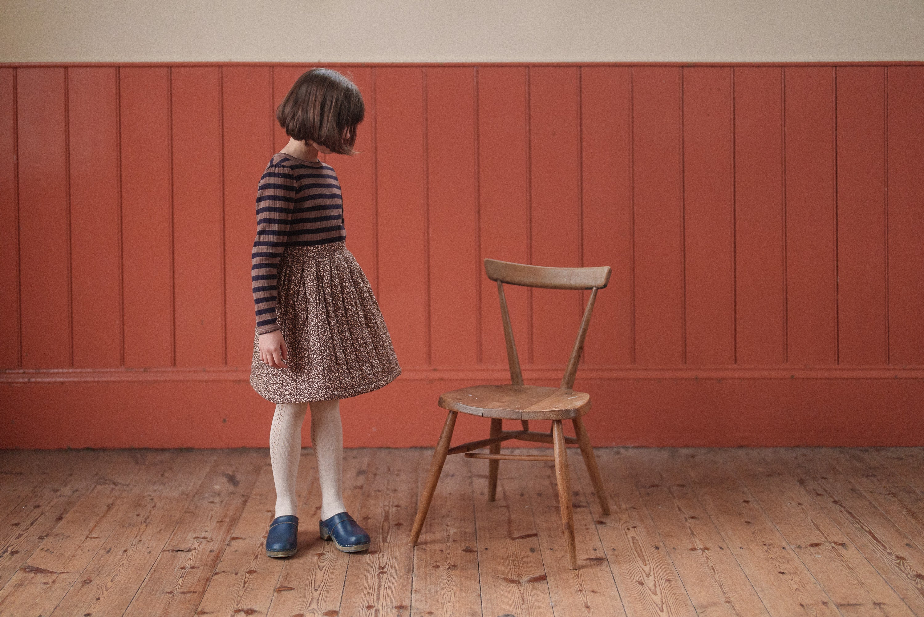 A young girl wearing a vintage-style striped navy and beige organic cotton slim fit t-shirt paired with a patterned skirt, white tights, and blue shoes, standing beside a wooden chair in a warm, softly lit room with natural wooden flooring and rust-colored paneled walls.