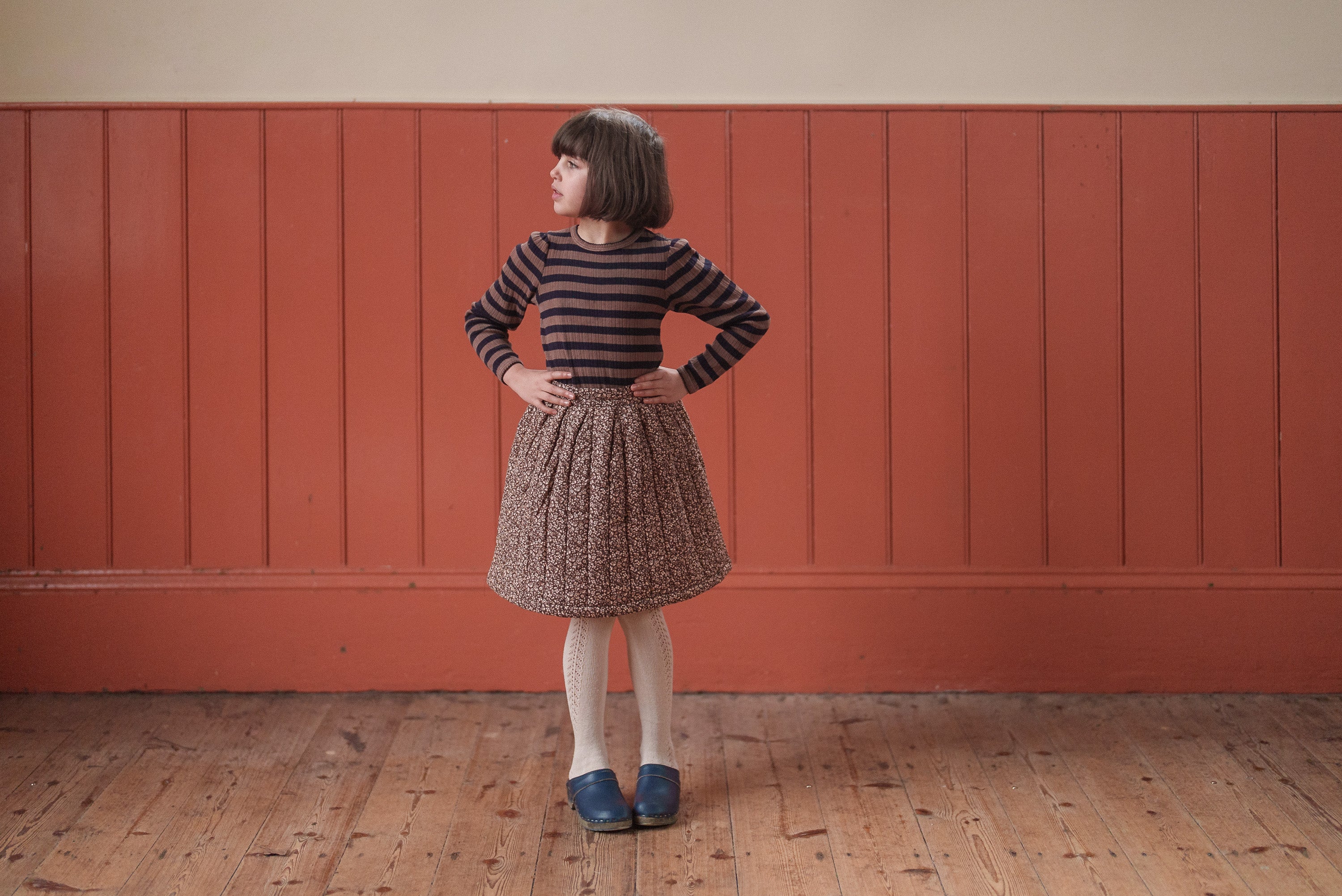 A young child wearing a navy and beige striped organic cotton slim fit T-shirt paired with a vintage-style gathered skirt, standing confidently on a wooden floor with hands on hips against a warm red paneled wall.